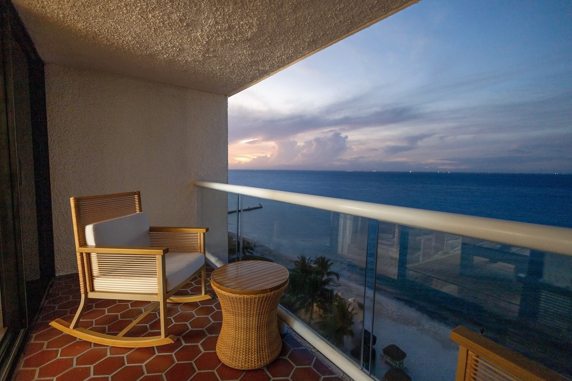 Oceanfront hotel balcony with rocking chair, table, and sunset view.
