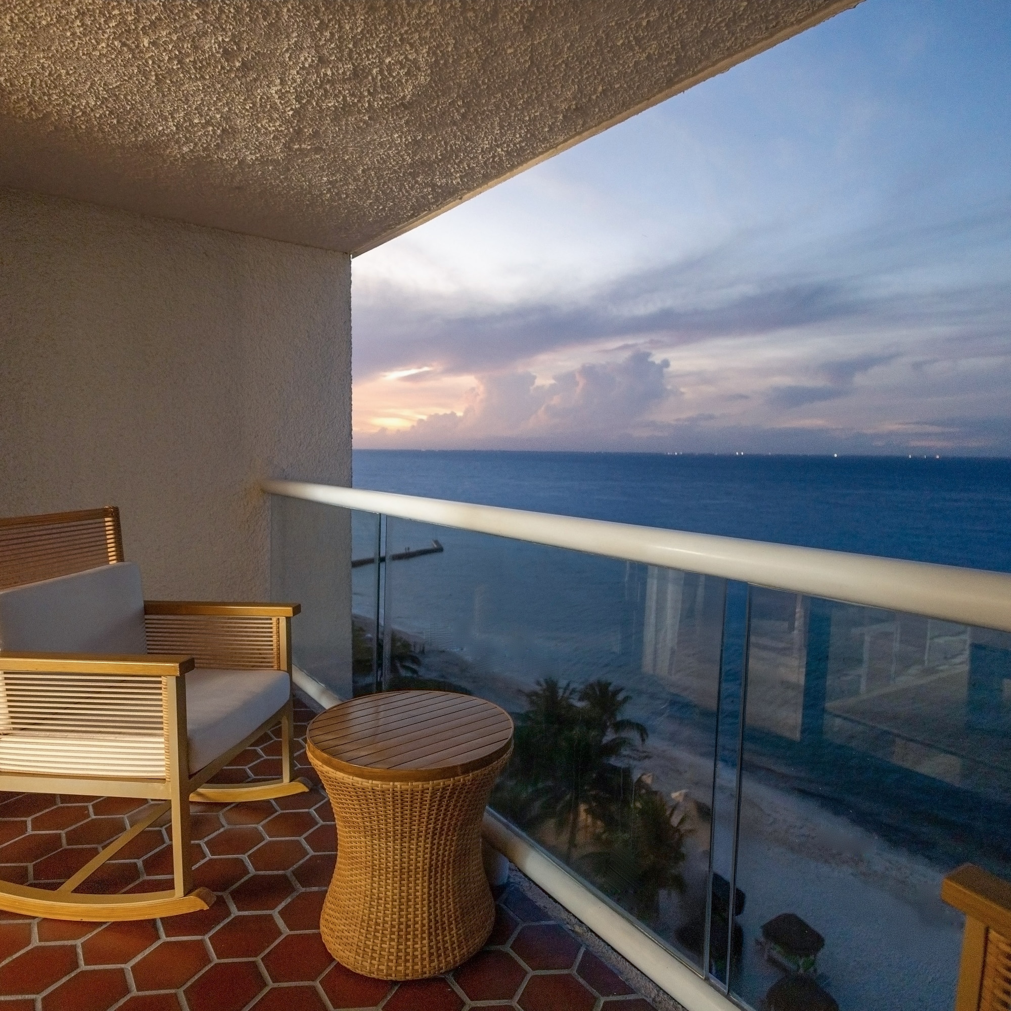 Oceanfront hotel balcony with rocking chair, table, and sunset view.