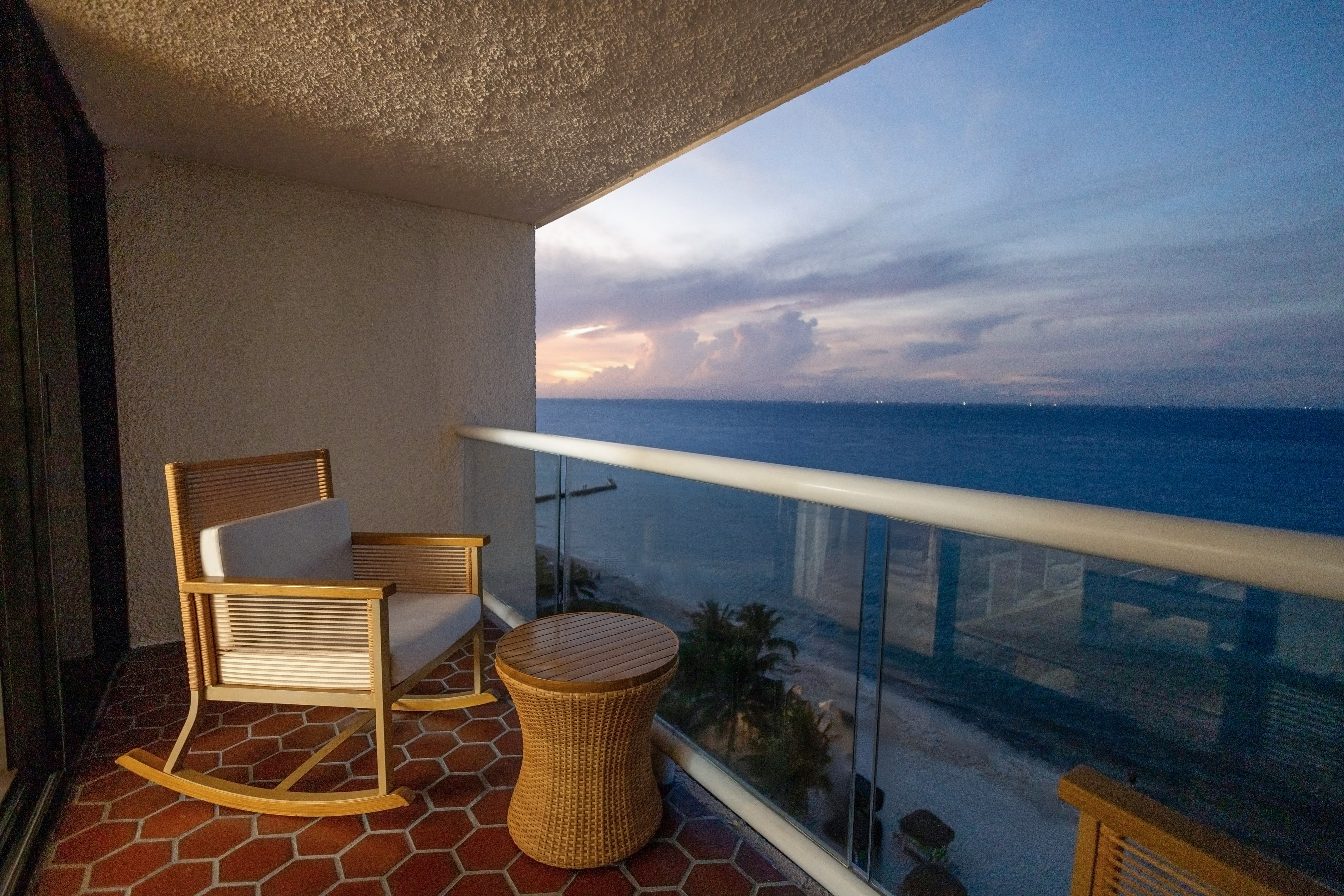 Oceanfront balcony with rocking chair, wicker table, sunset ocean view.