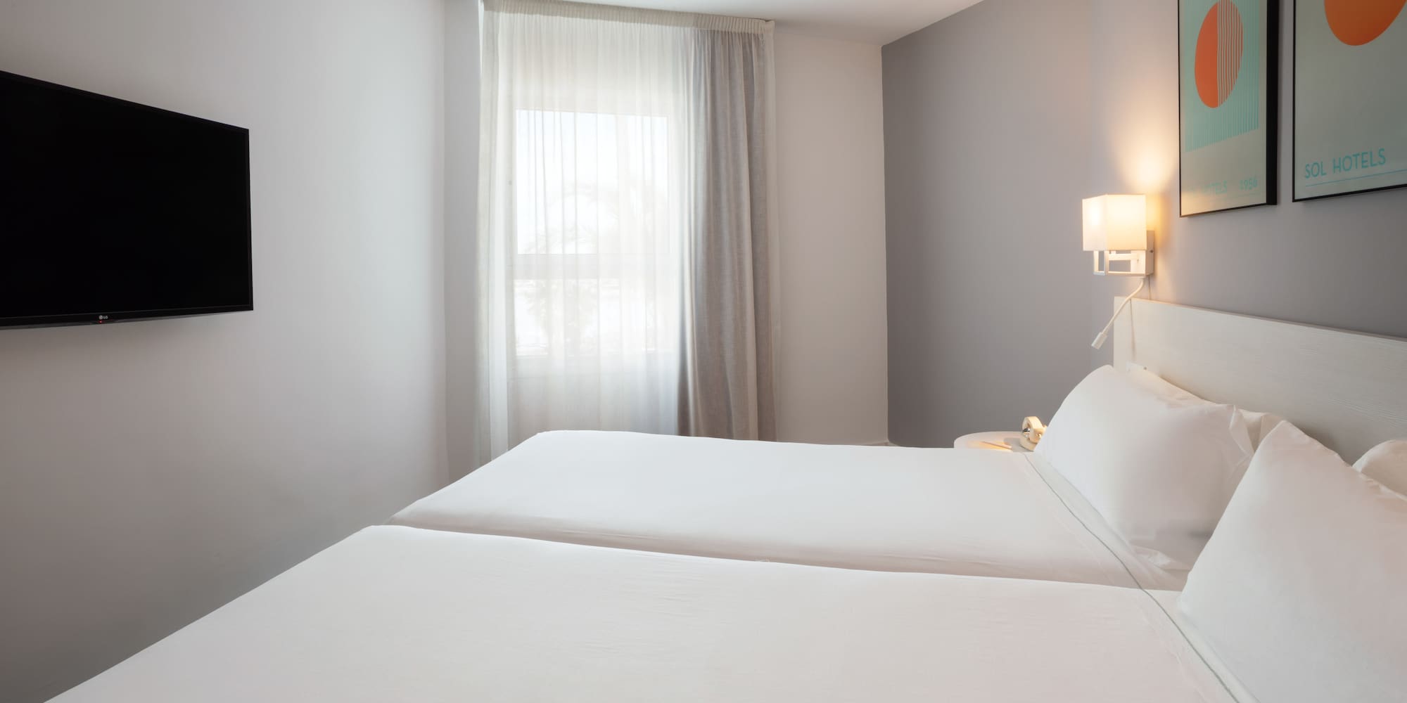 Modern hotel room with twin beds, wall-mounted TV, and window.