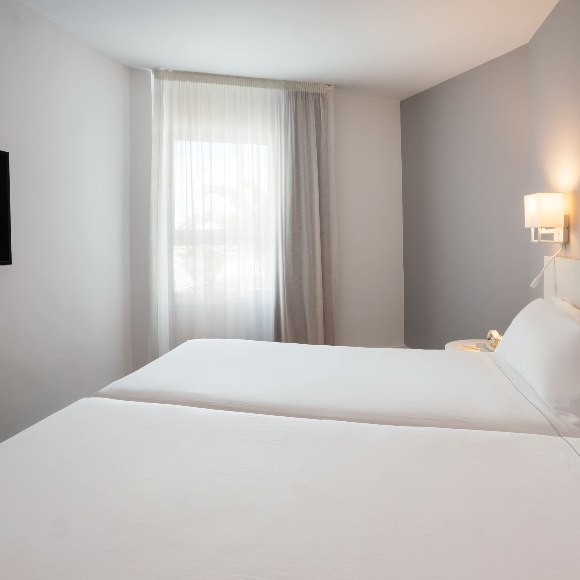 Modern hotel room with twin beds, wall-mounted TV, and window.