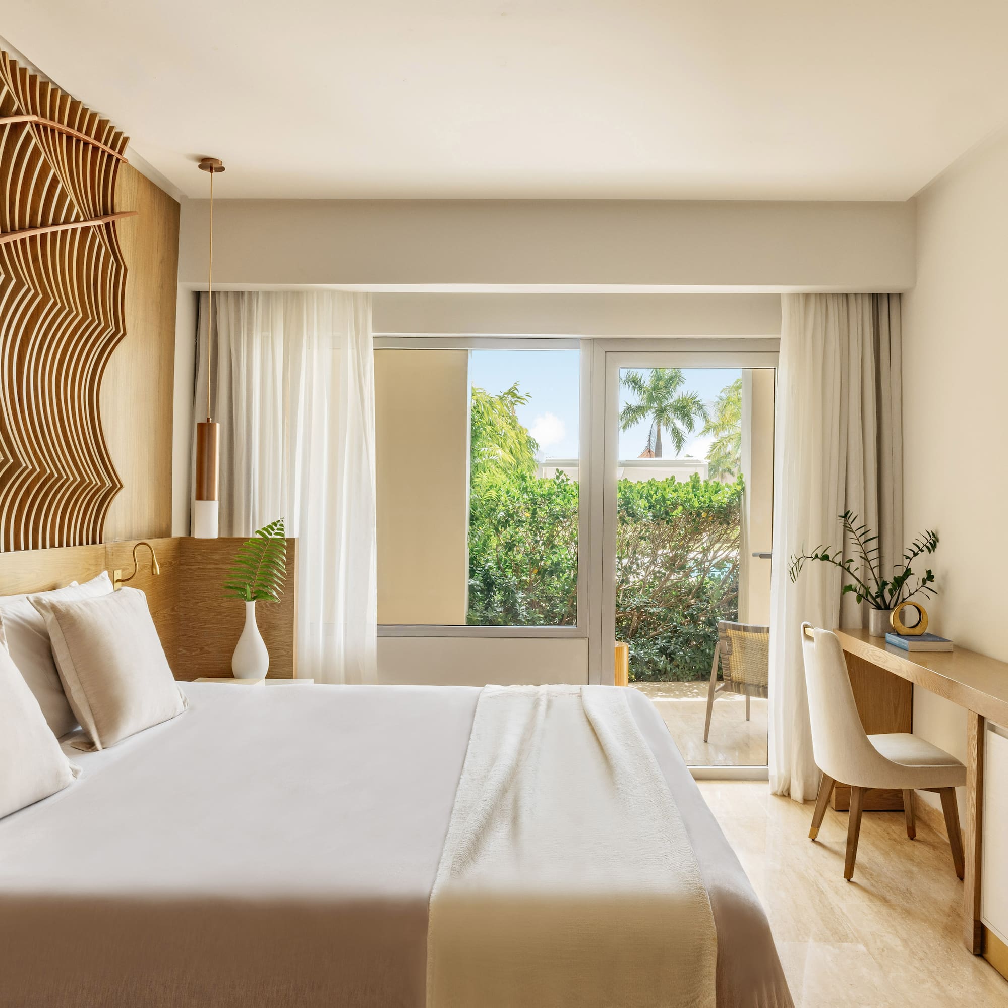 Elegant hotel bedroom with wooden headboard, bed, and tropical balcony view.