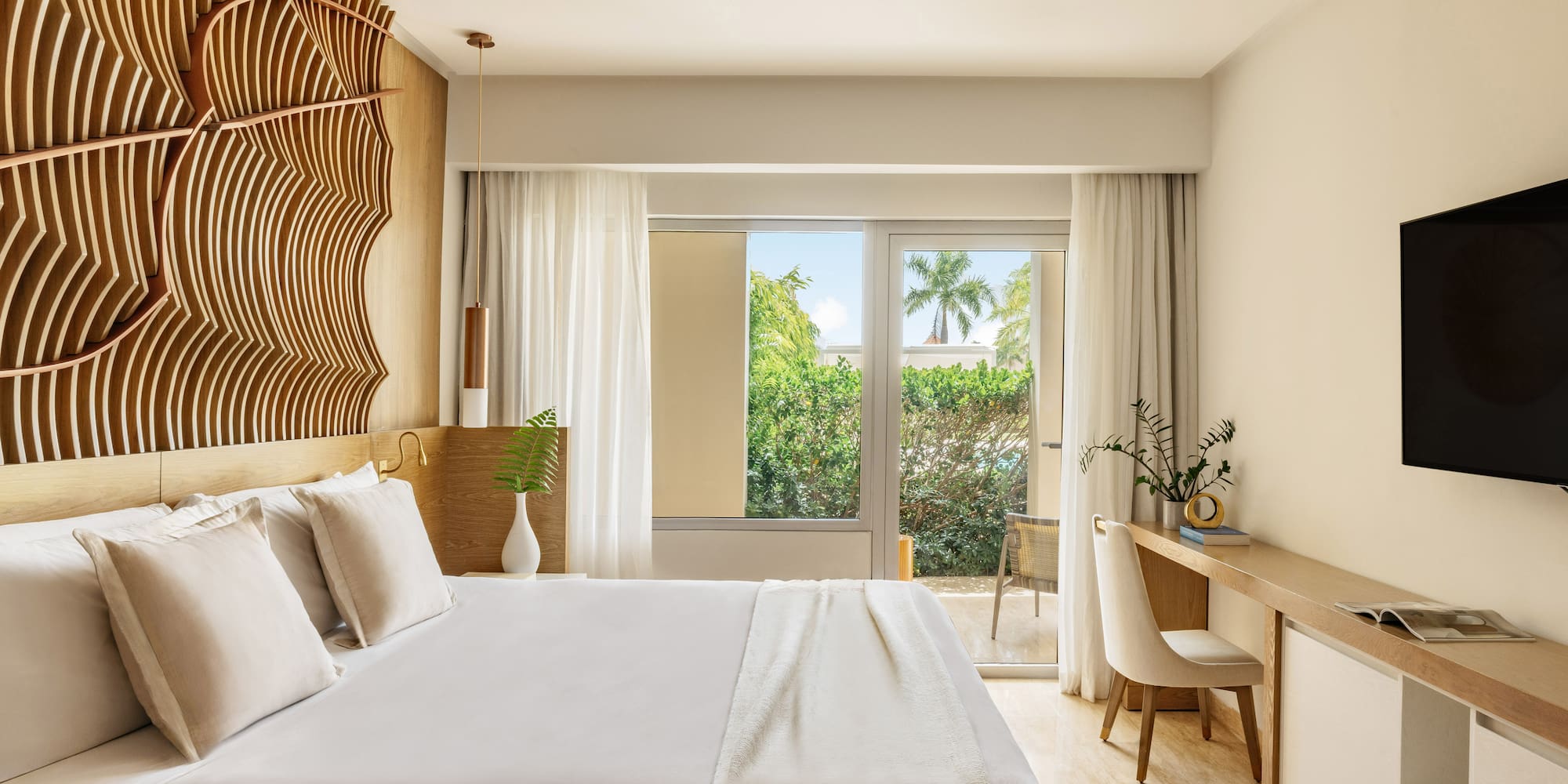Elegant hotel bedroom with wooden headboard, bed, and tropical balcony view.