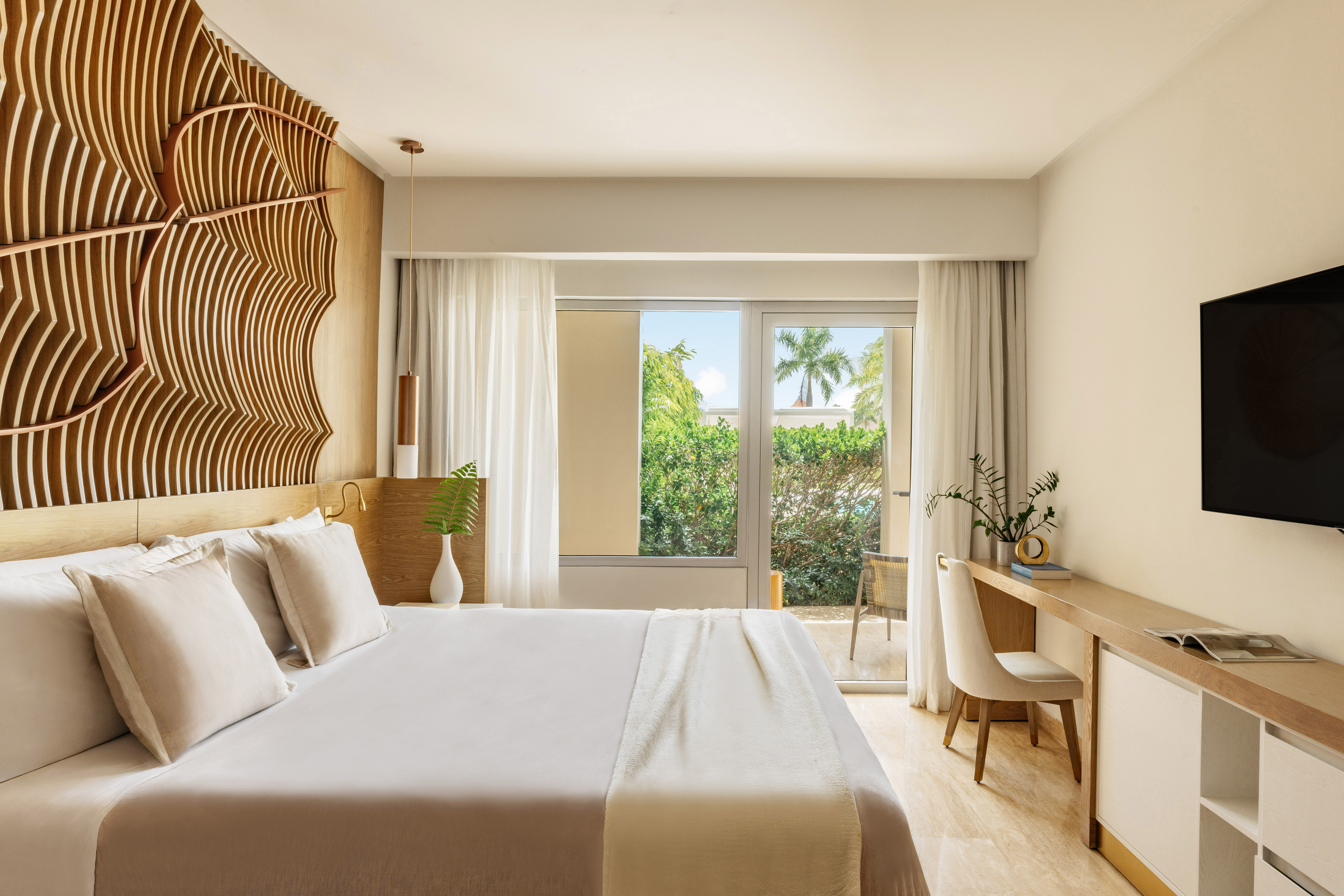 Elegant hotel bedroom with wooden headboard, bed, and tropical balcony view.