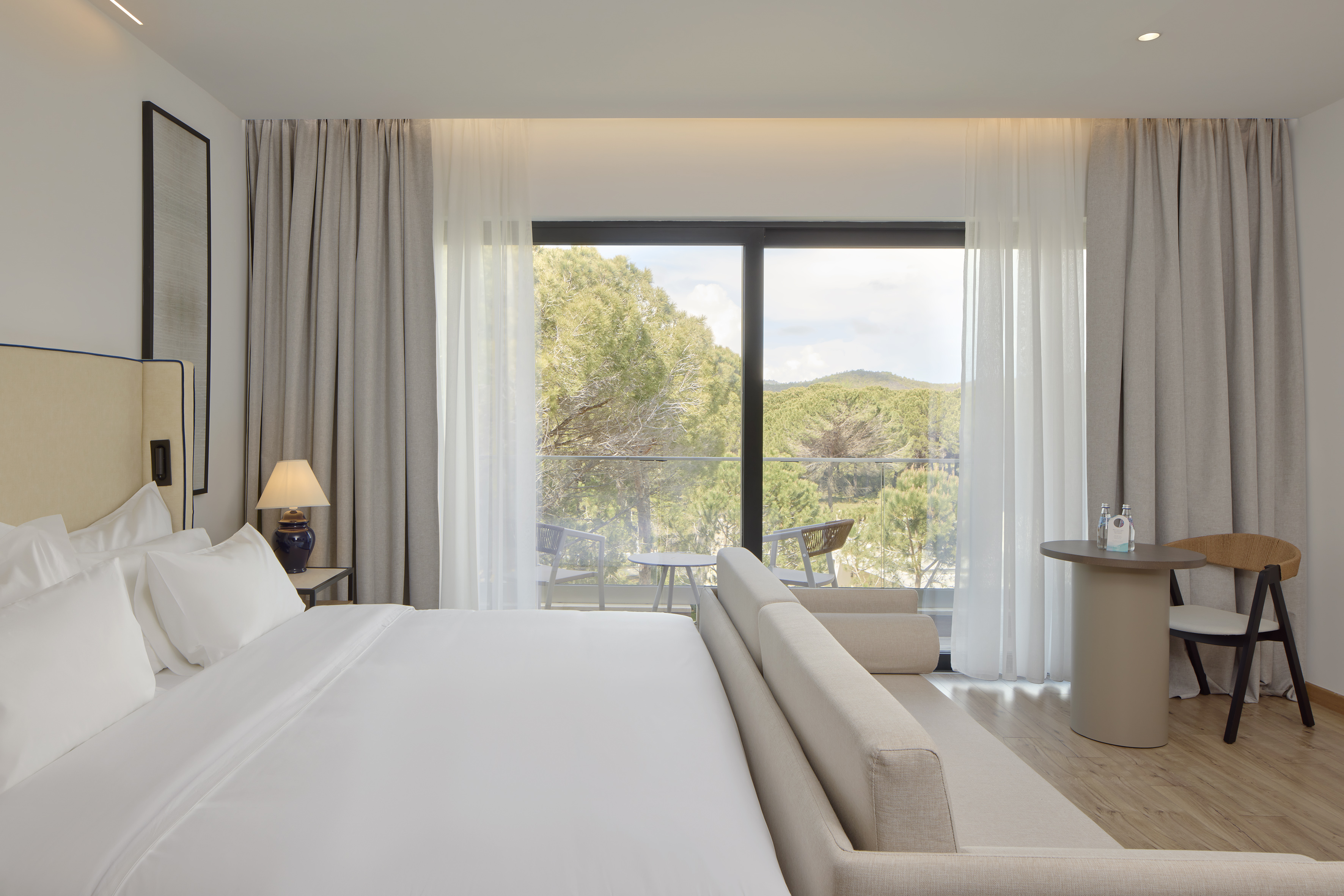 Hotel bedroom with white bed, balcony access, and scenic forest view.