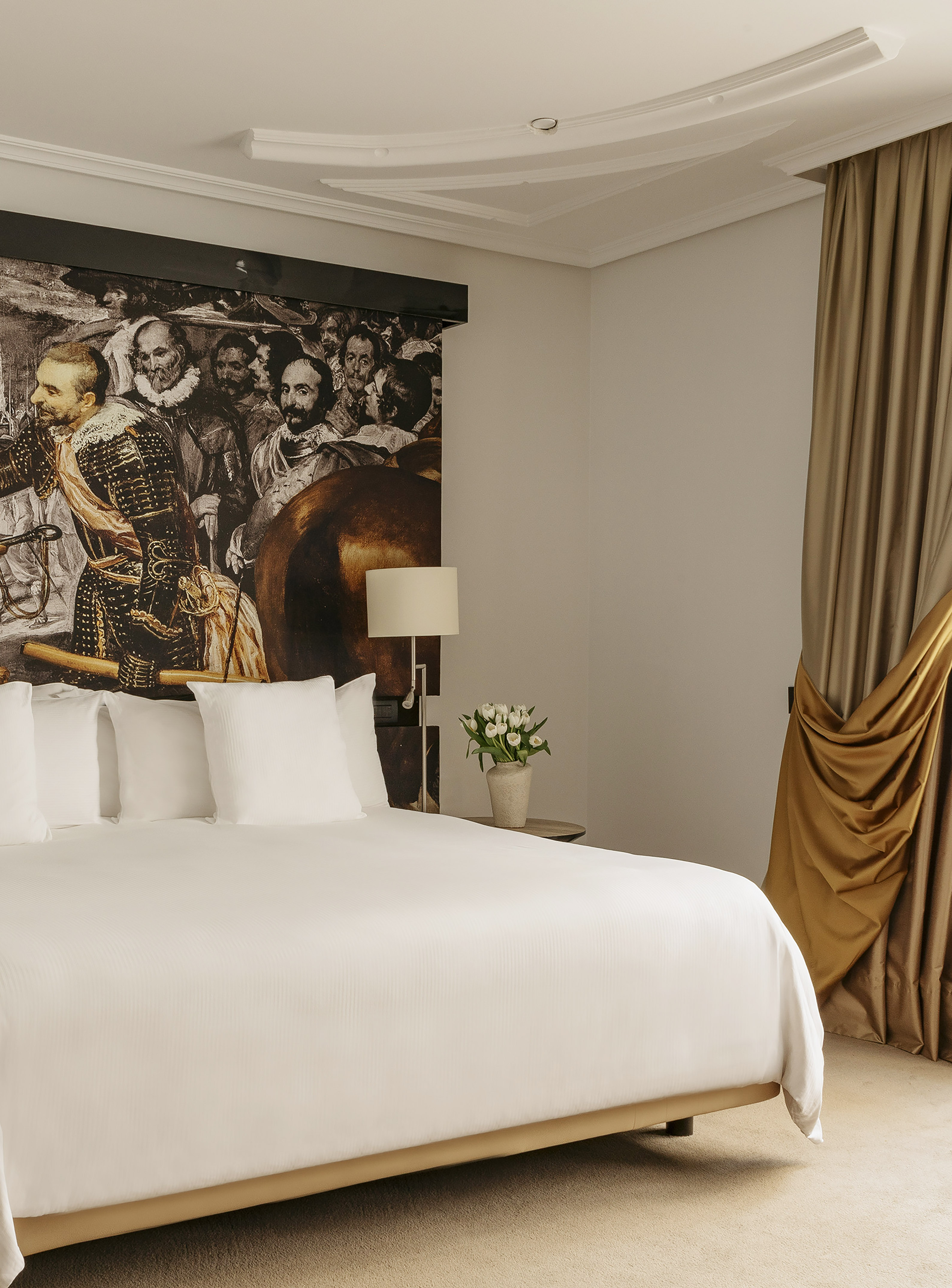 Hotel room with large bed, historical art mural headboard, white tulips, and gold curtains.