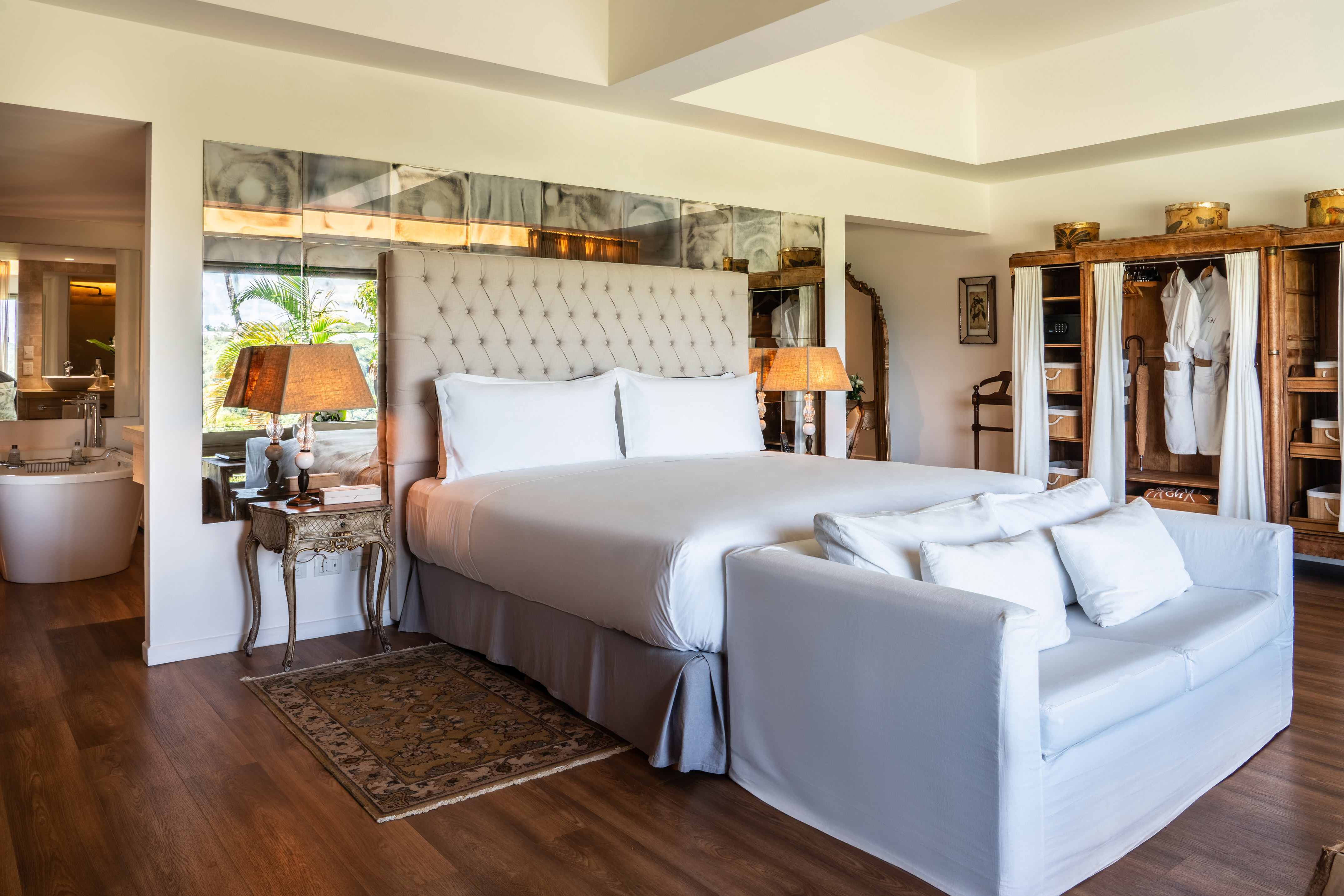 Luxury hotel suite with tufted bed, mirrored wall, wooden armoire, and spa bath.