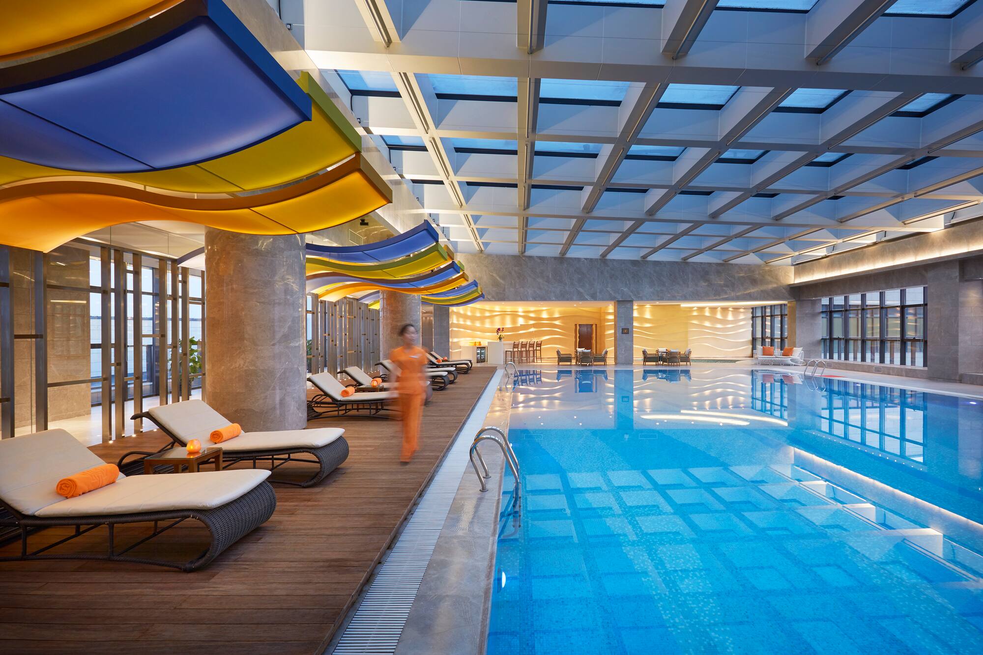 Indoor hotel pool with colorful wavy ceiling, skylight, and loungers.