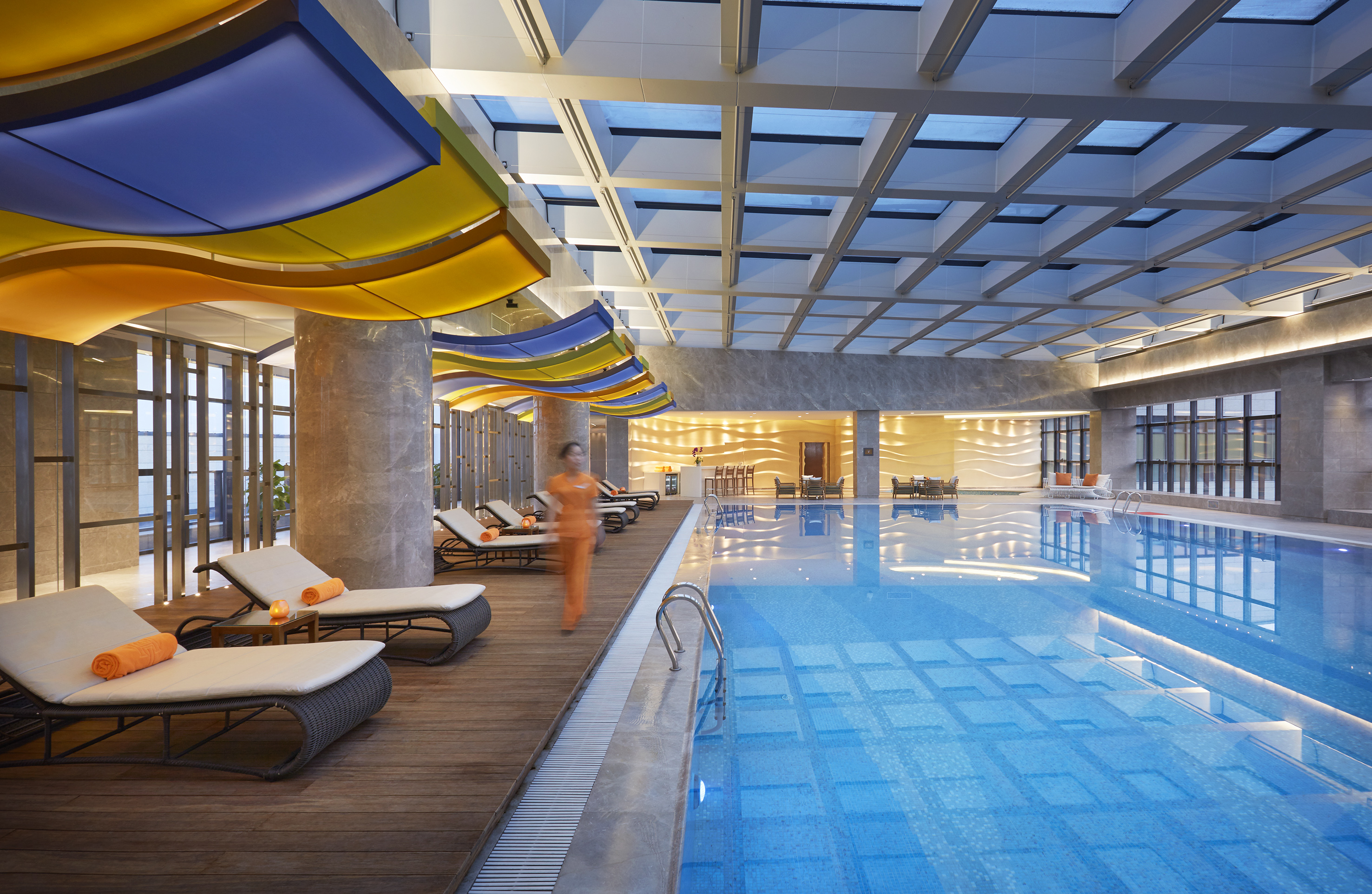Indoor hotel pool with colorful wavy ceiling, skylight, and loungers.