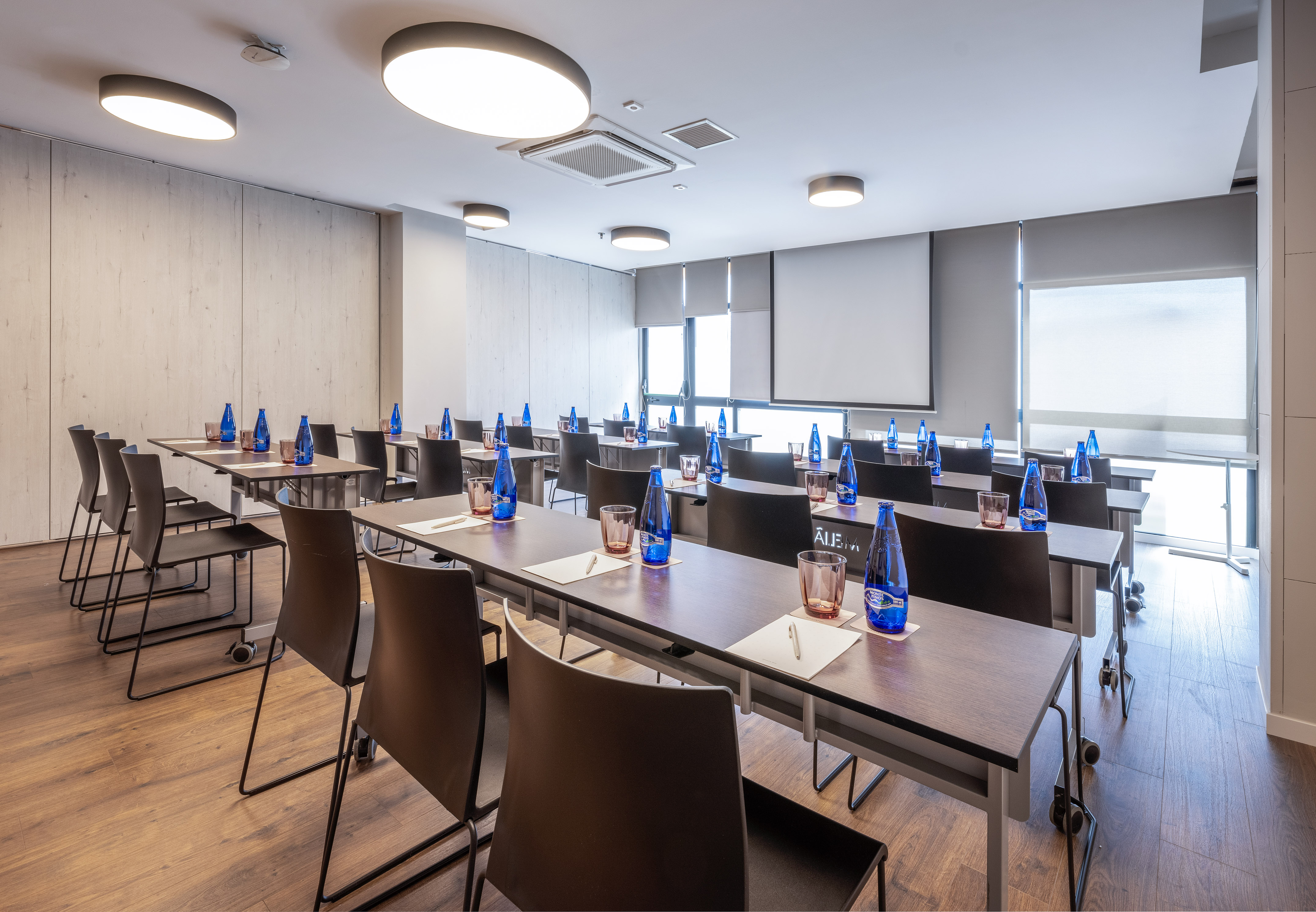 Modern meeting room with classroom setup, tables, chairs, water, projector screen.