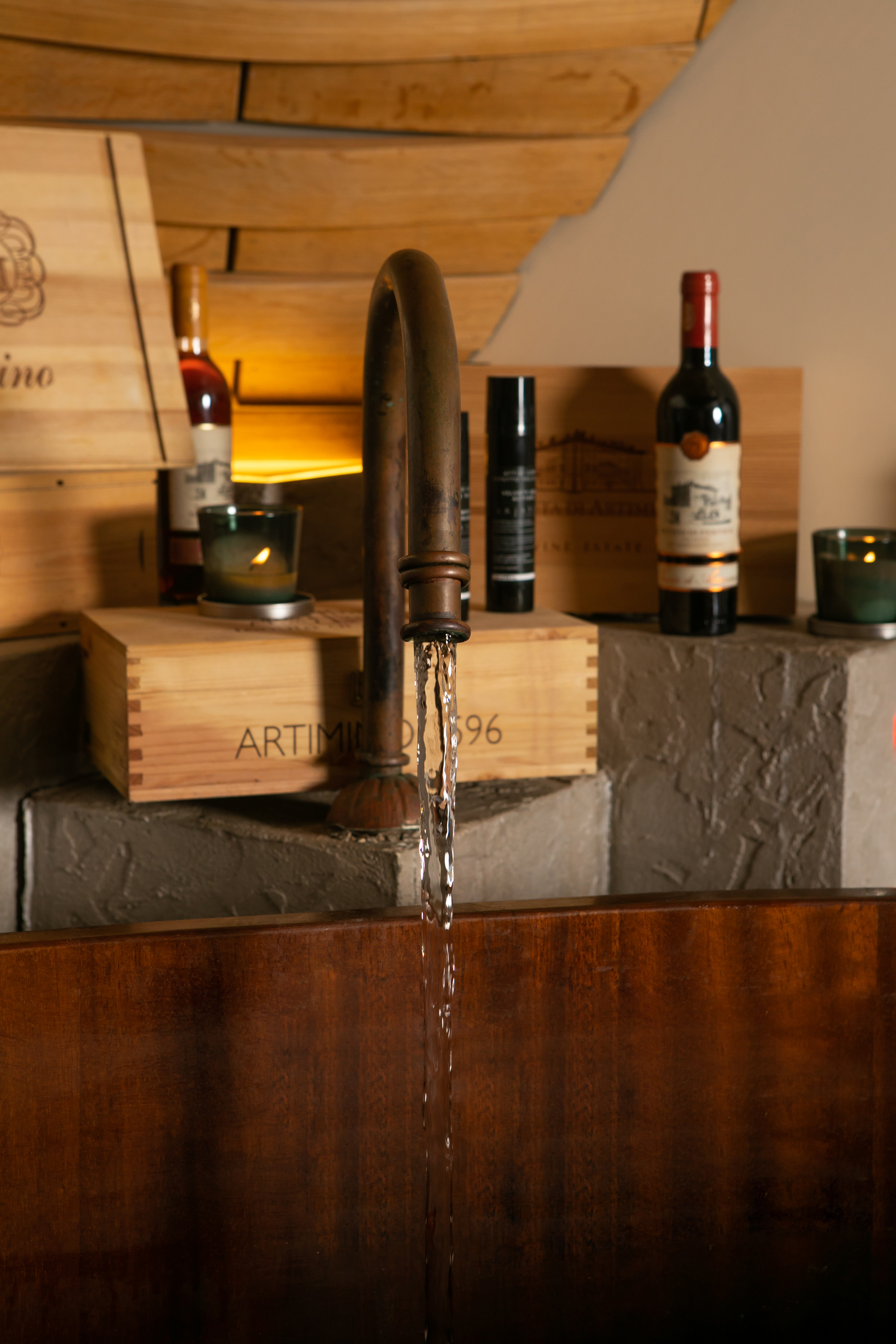 Rustic bronze faucet pouring water into wooden basin, surrounded by wine crates.