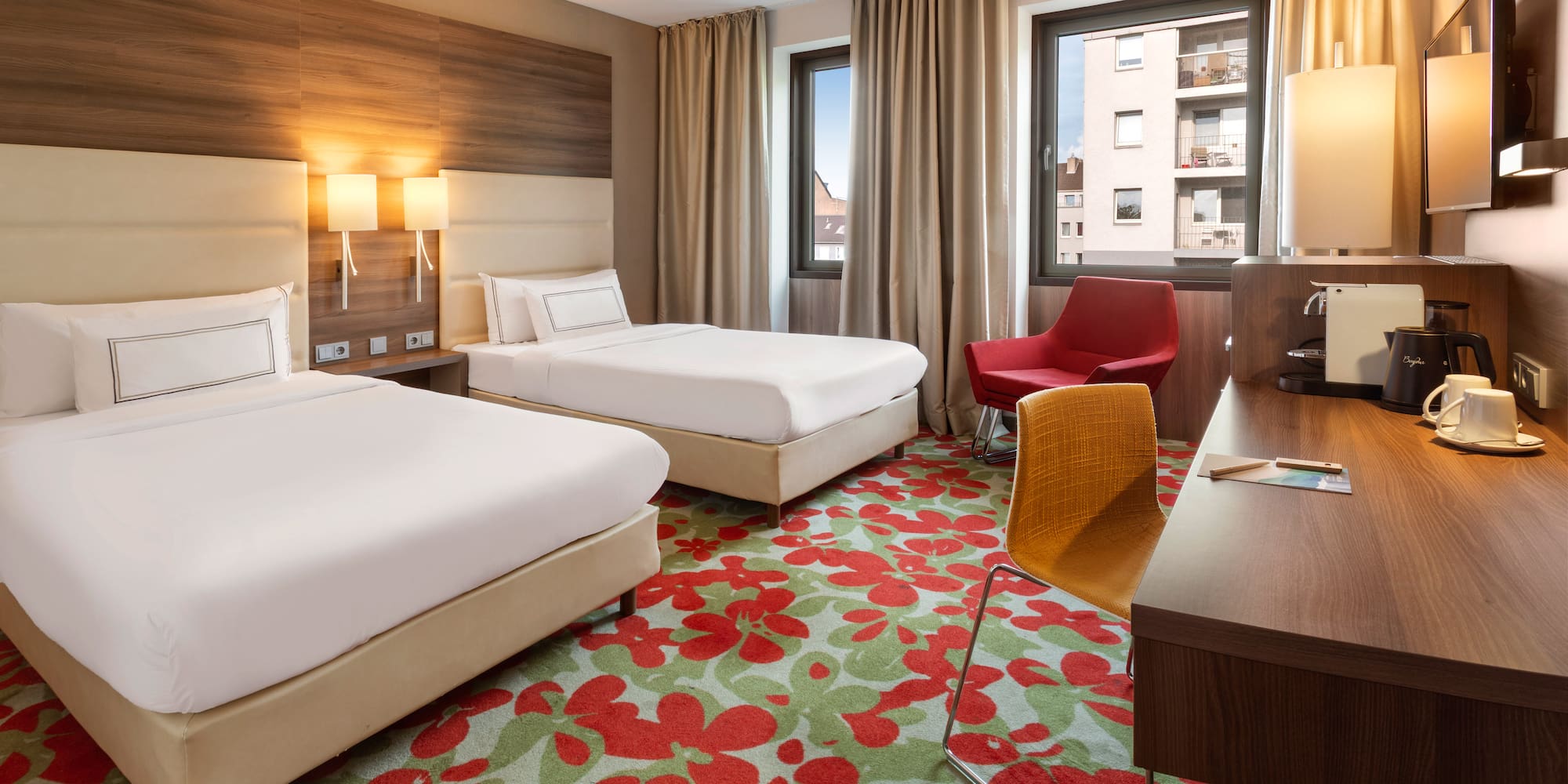 Modern hotel room with twin beds, floral carpet, work desk, and armchair.