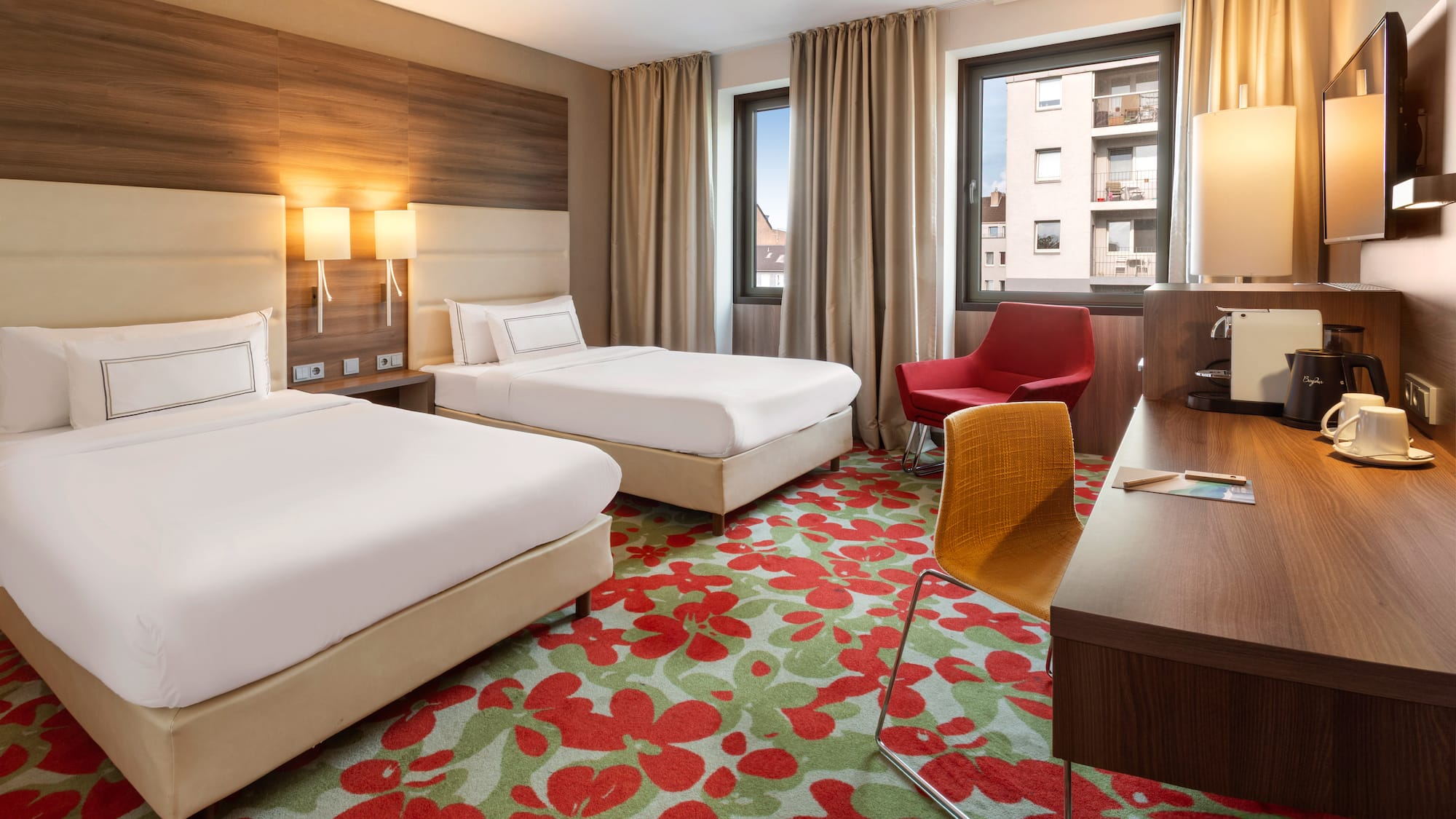 Modern hotel room with twin beds, floral carpet, work desk, and armchair.