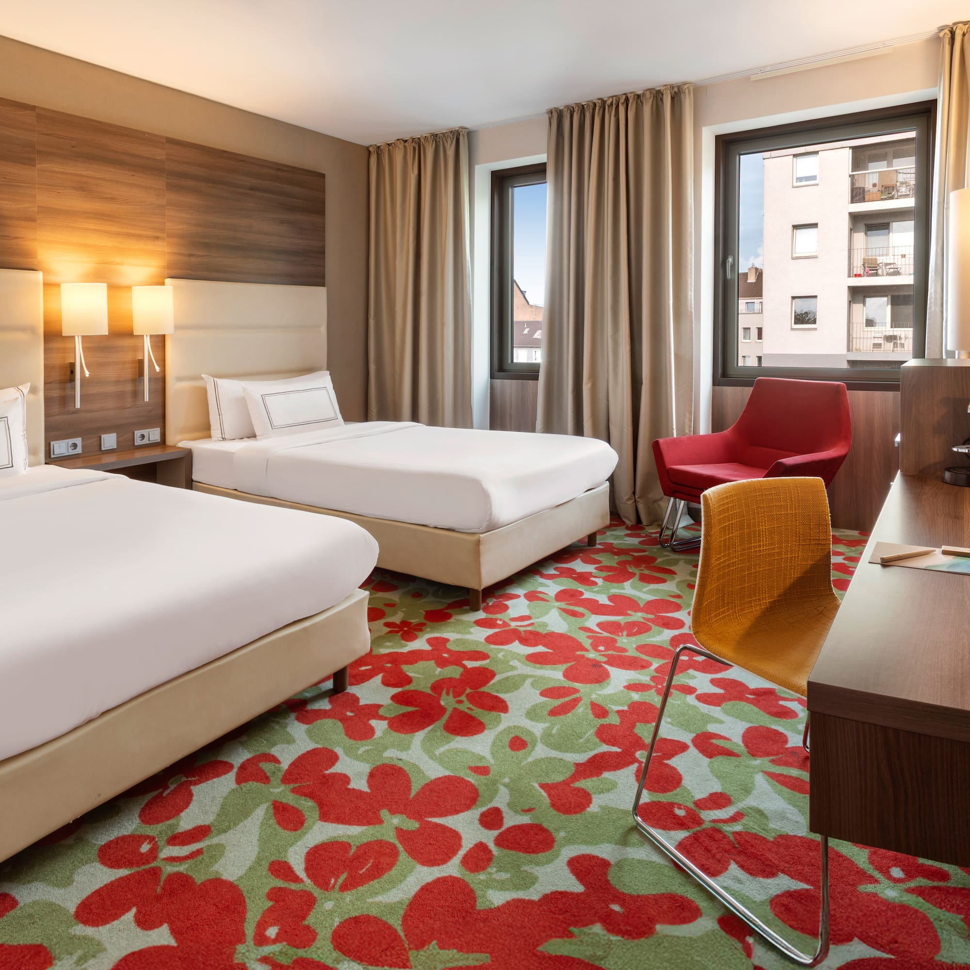Modern hotel room with twin beds, floral carpet, work desk, and armchair.