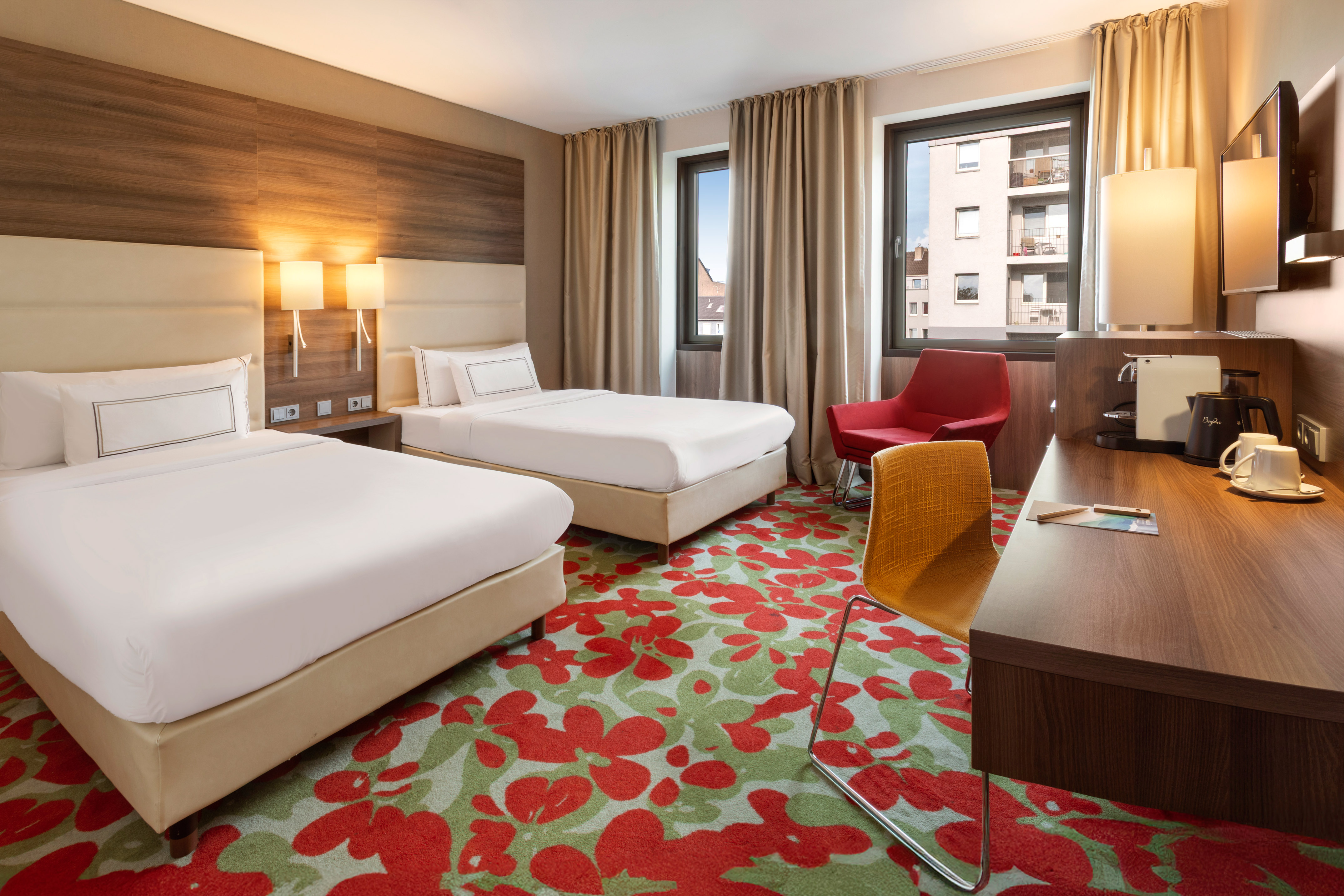 Modern hotel room with twin beds, floral carpet, work desk, and armchair.