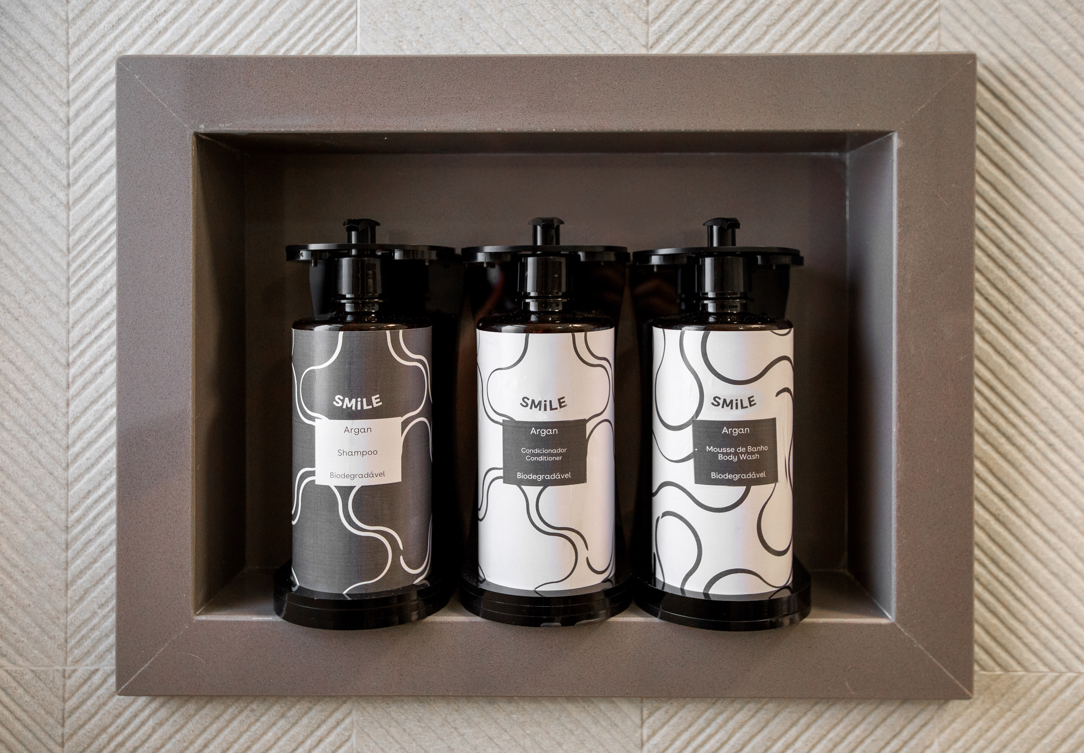 Three "SMILE" Argan shampoo, conditioner, body wash bottles in a shower niche.