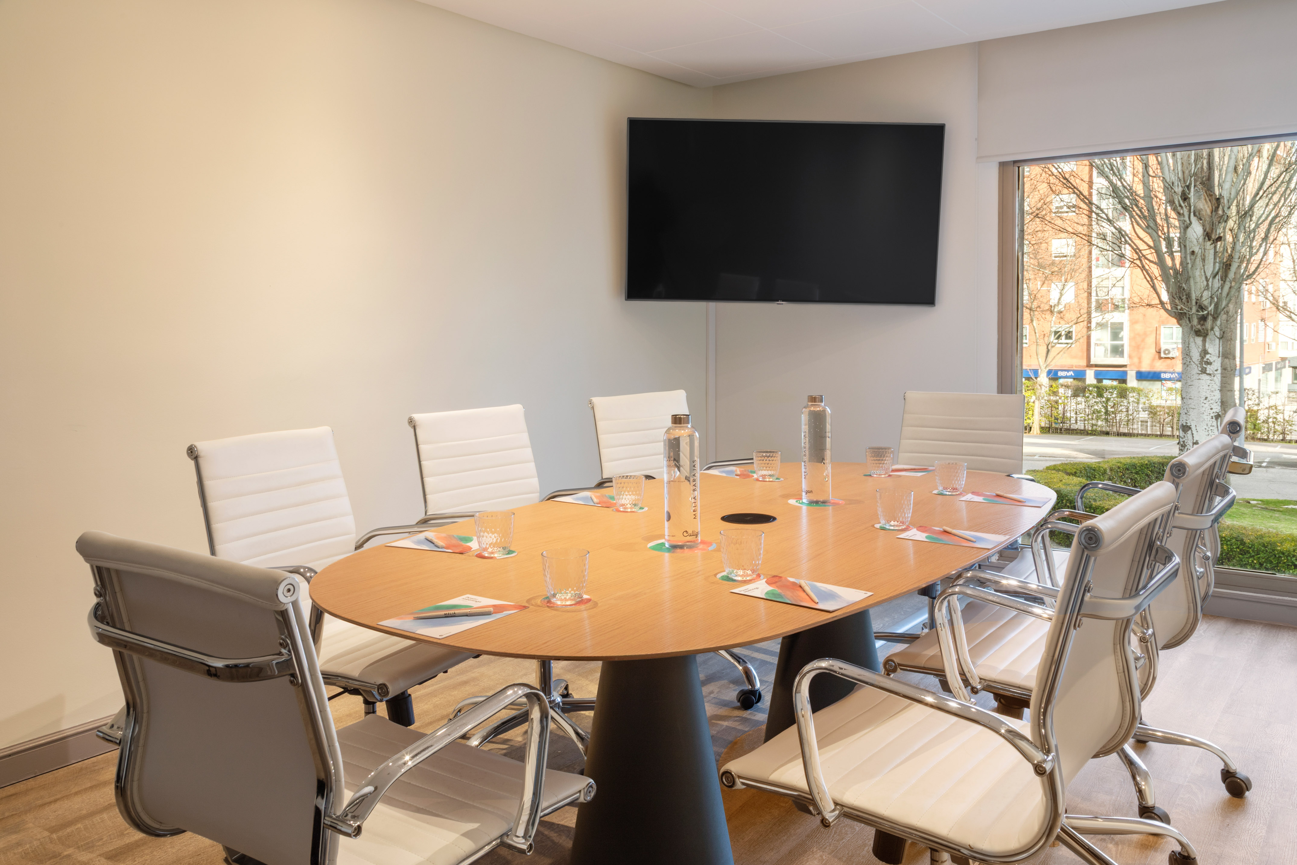 Modern conference room with oval wood table, white chairs, TV, and cityscape view.