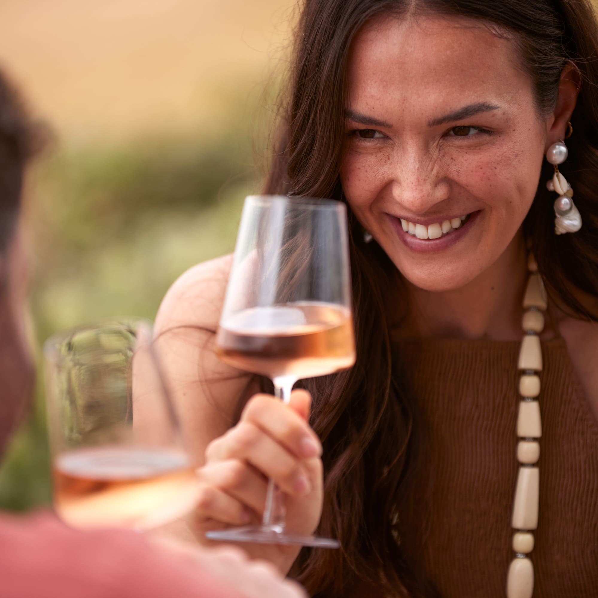 Smiling woman holding rosé wine, toasting with companion outdoors.