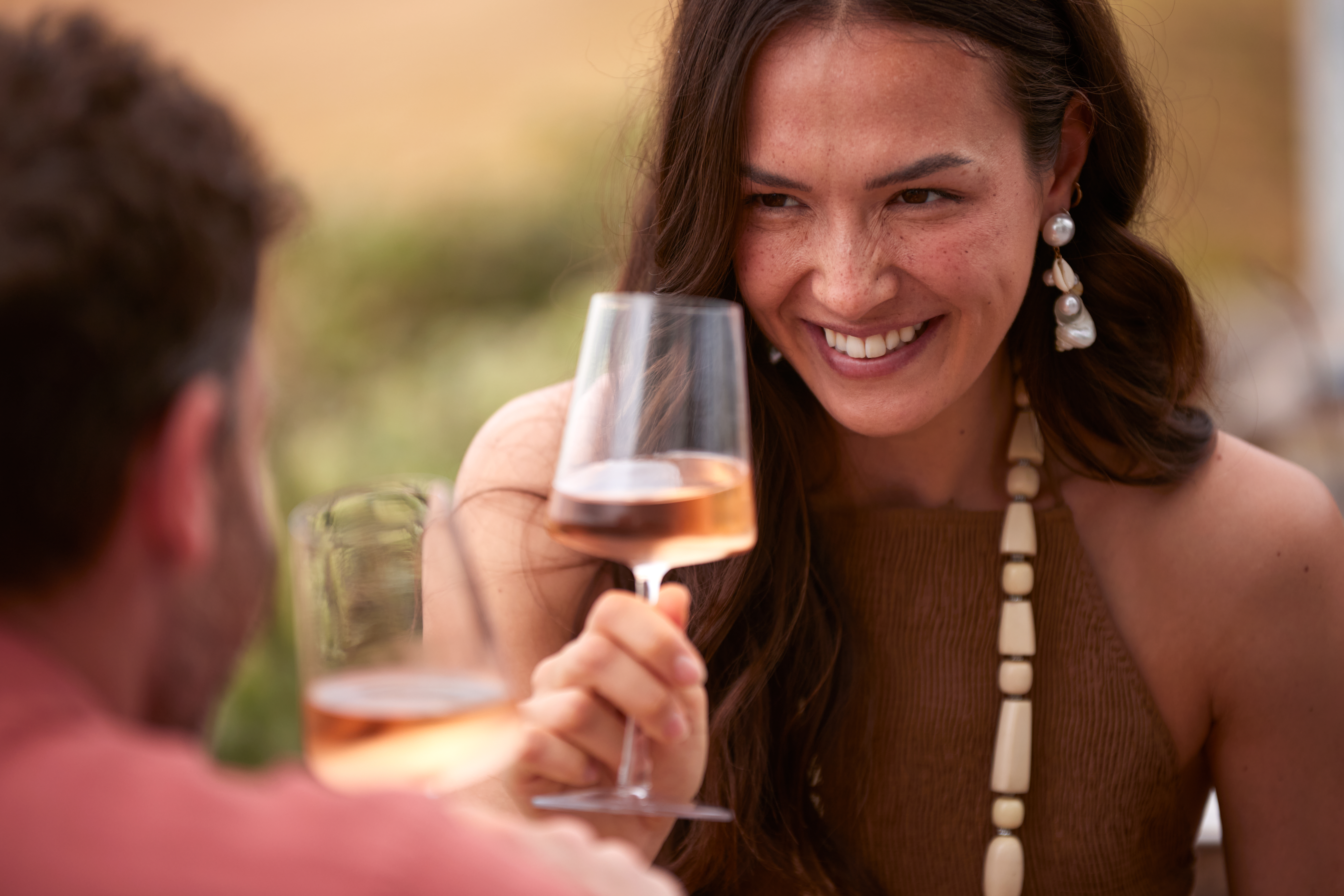 Smiling woman holding rosé wine, toasting with companion outdoors.