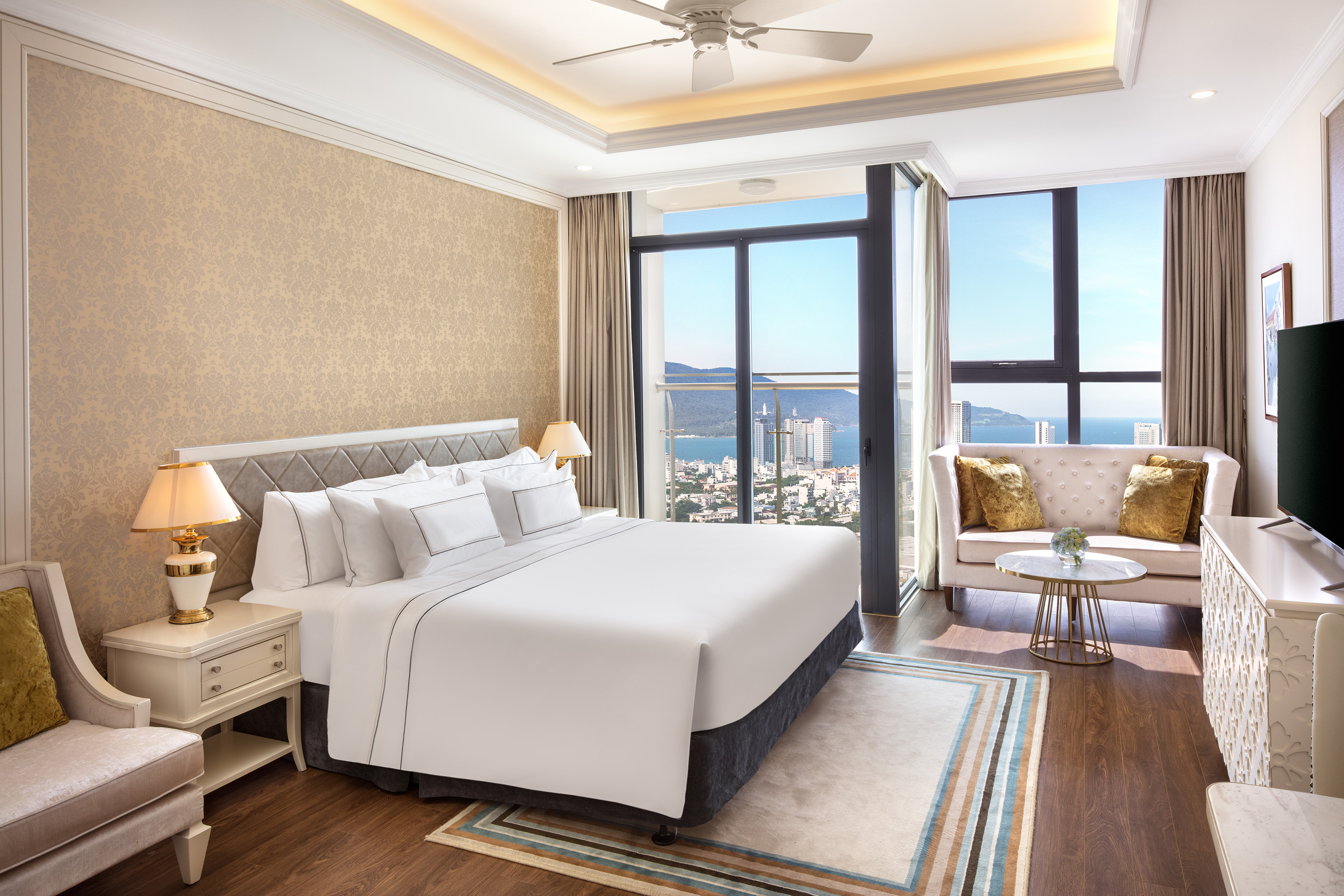 Luxury hotel room with king bed, sofa, and panoramic city-ocean view.