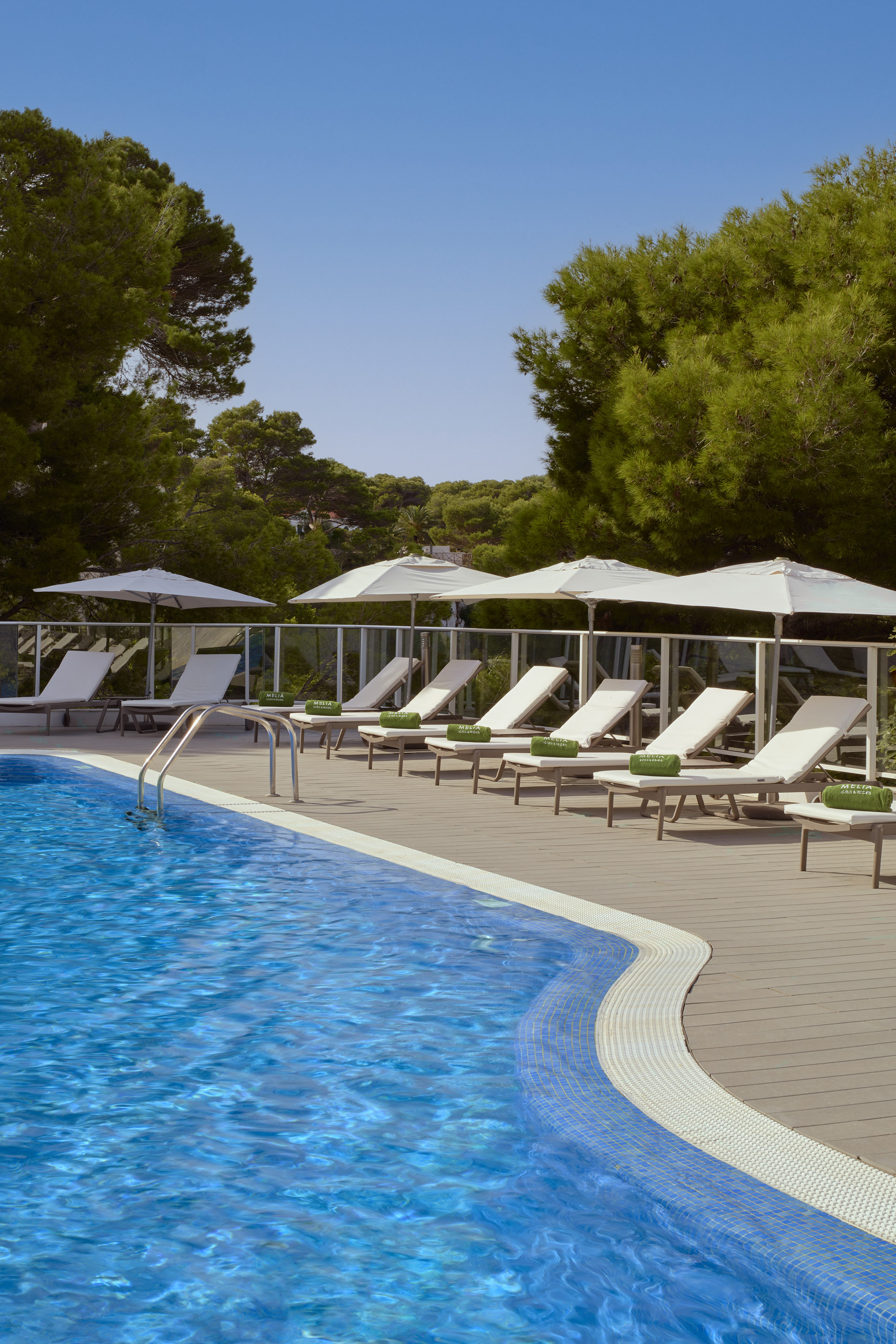 Resort swimming pool with blue water, sun loungers, and green pine trees.