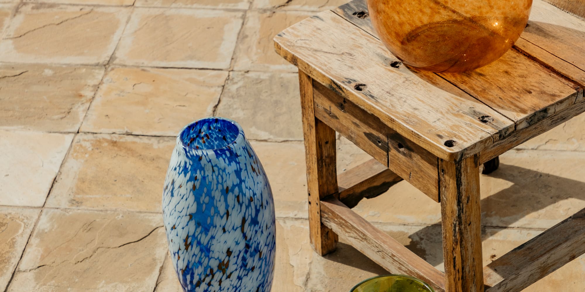 Speckled orange, blue, green glass vases on rustic tiles and wooden stool.