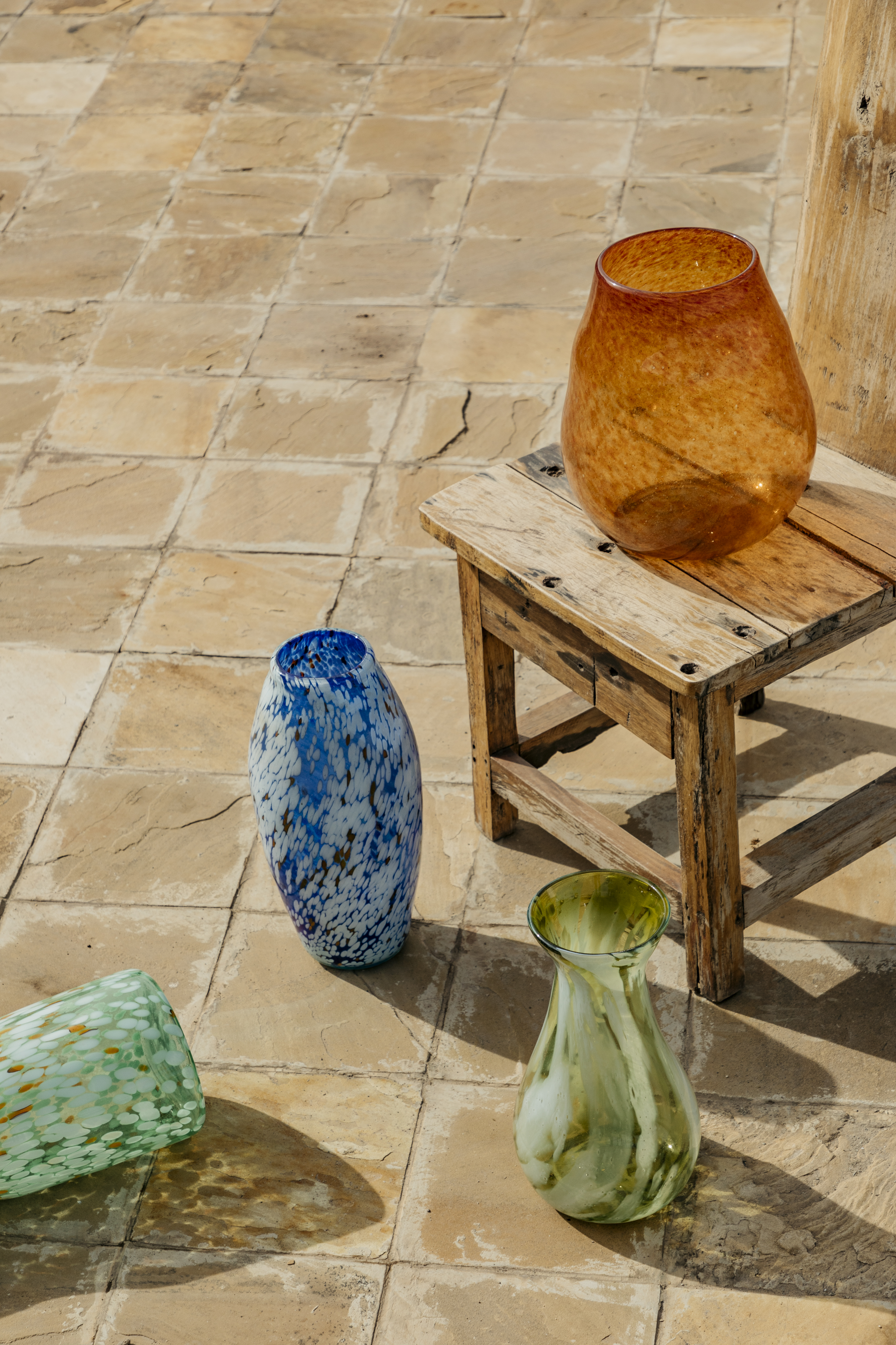 Speckled orange, blue, green glass vases on rustic tiles and wooden stool.