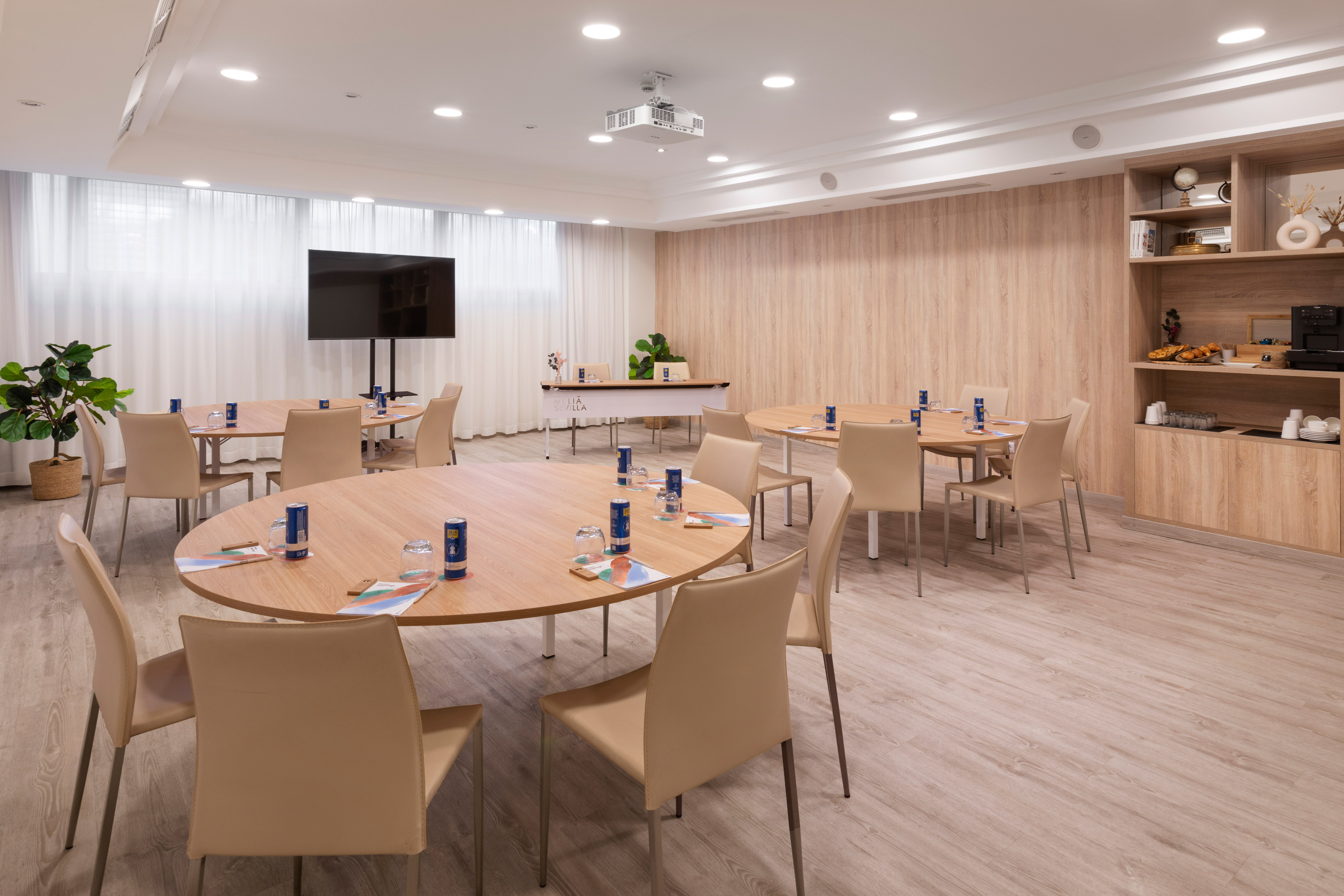 Modern meeting room with round tables, TV, projector, and refreshment station.