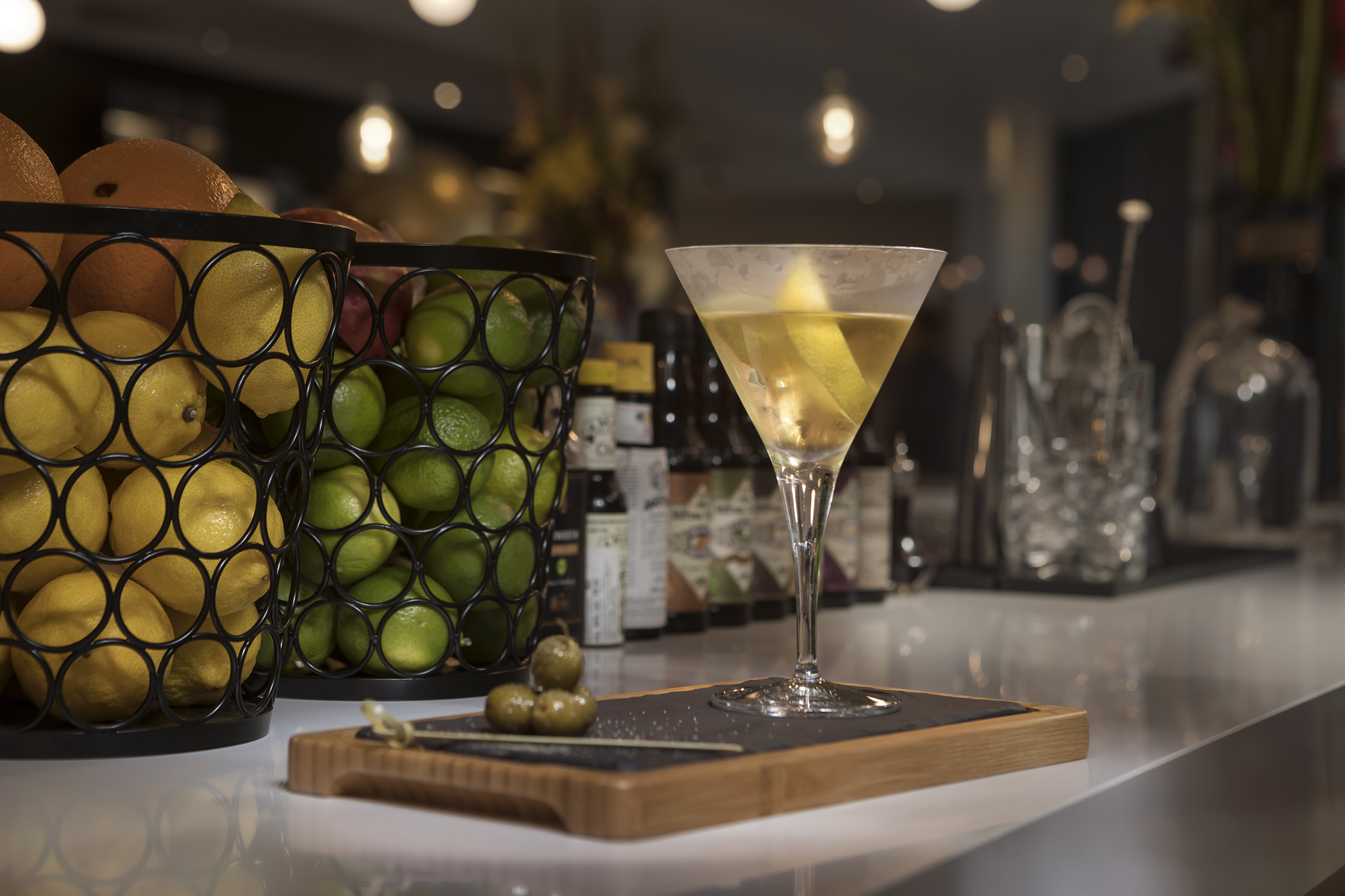 Martini with lemon twist and olives at a bar, with fresh lemons and limes.