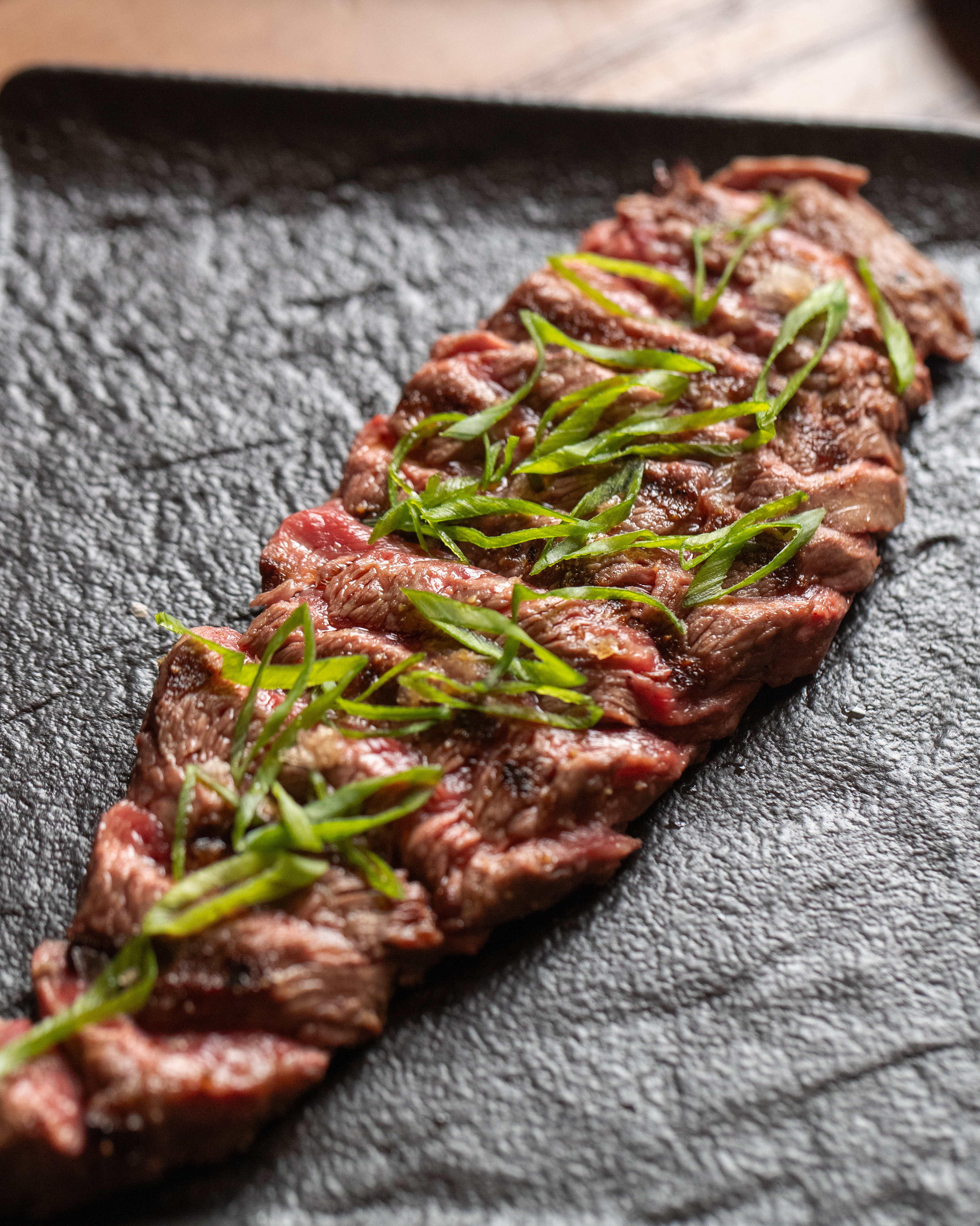 Sliced grilled steak topped with green onions on a dark textured plate.