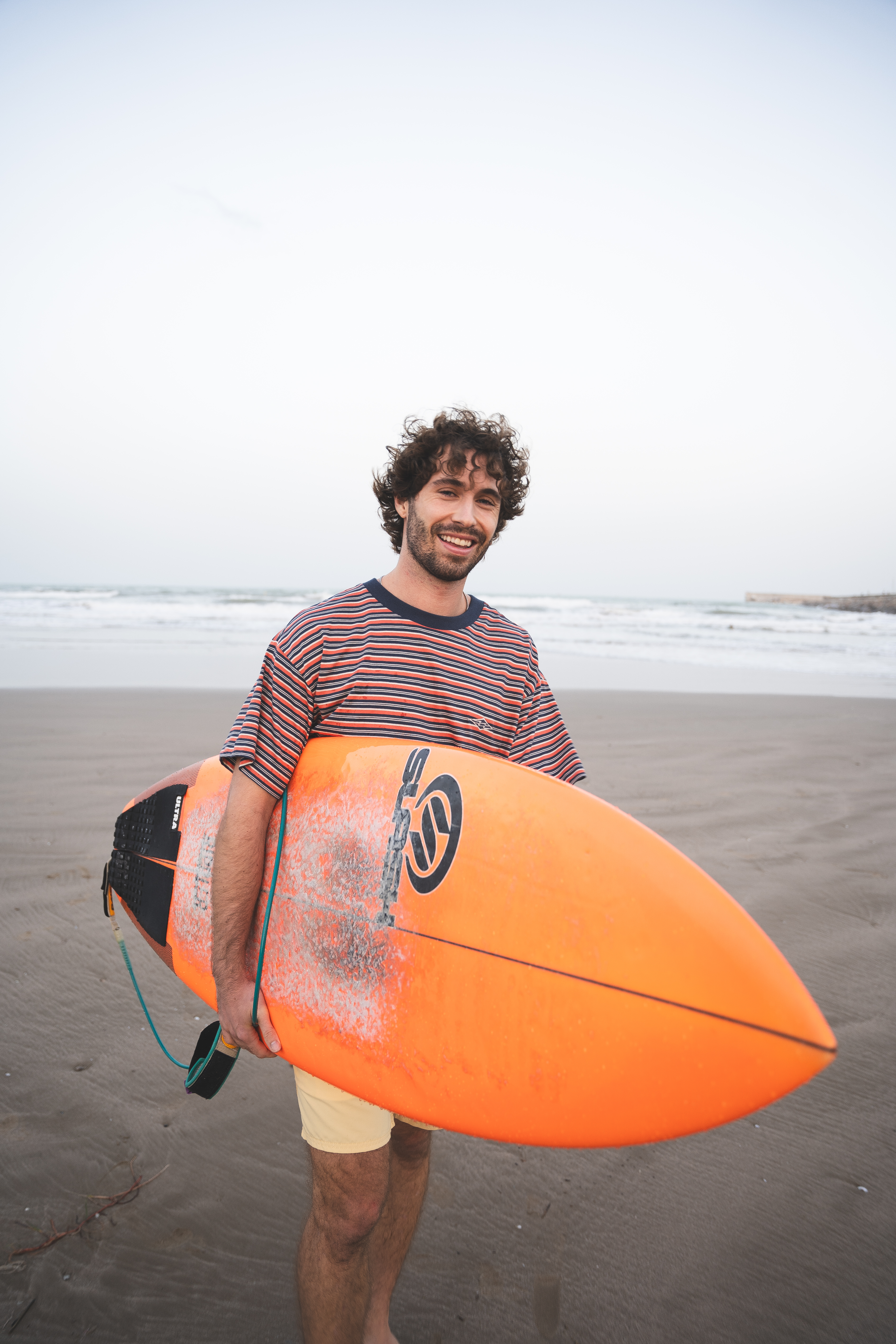 Happy surfer with orange board on sandy beach near ocean waves.