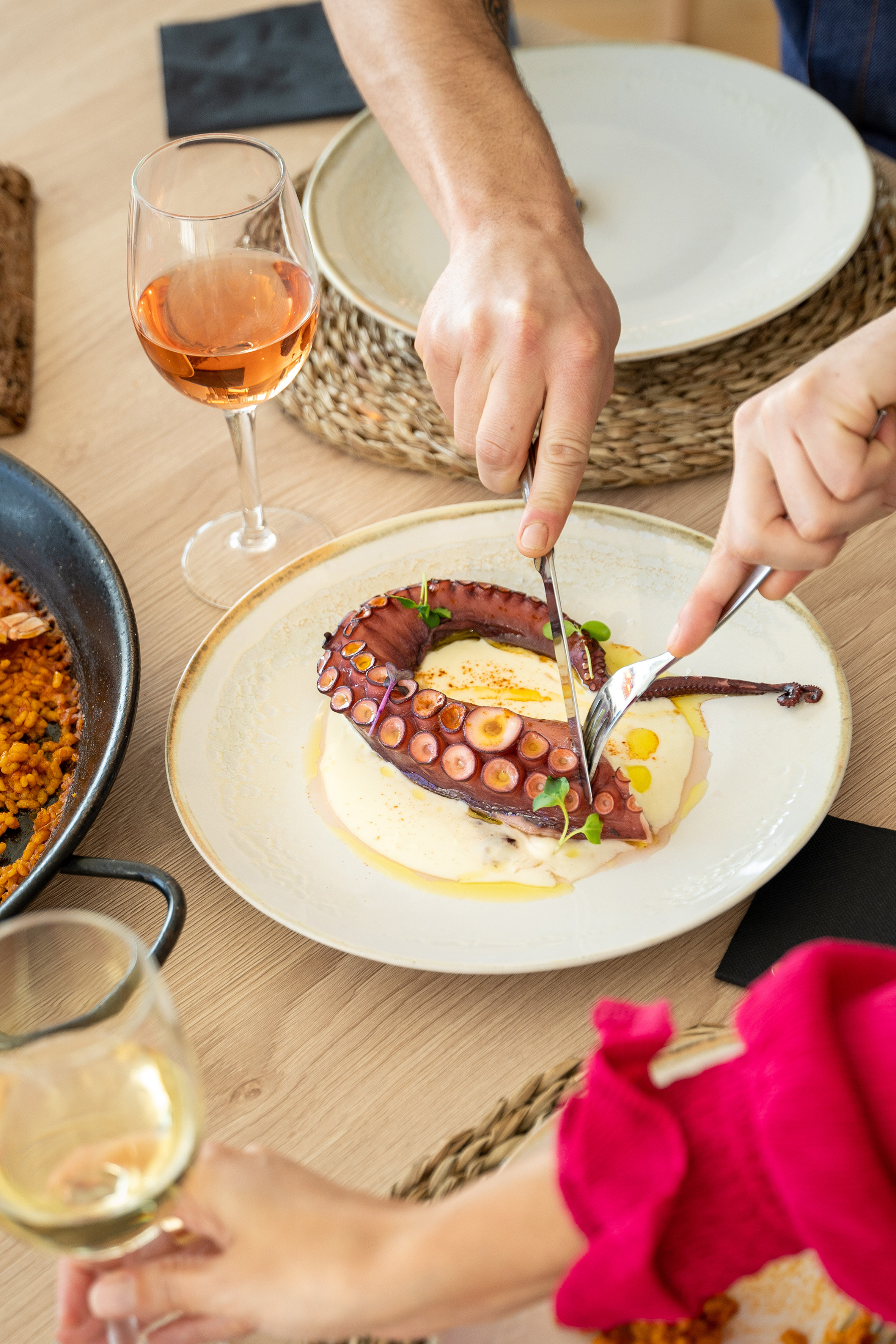 Hands cutting grilled octopus tentacle, served with puree, wine, and paella.