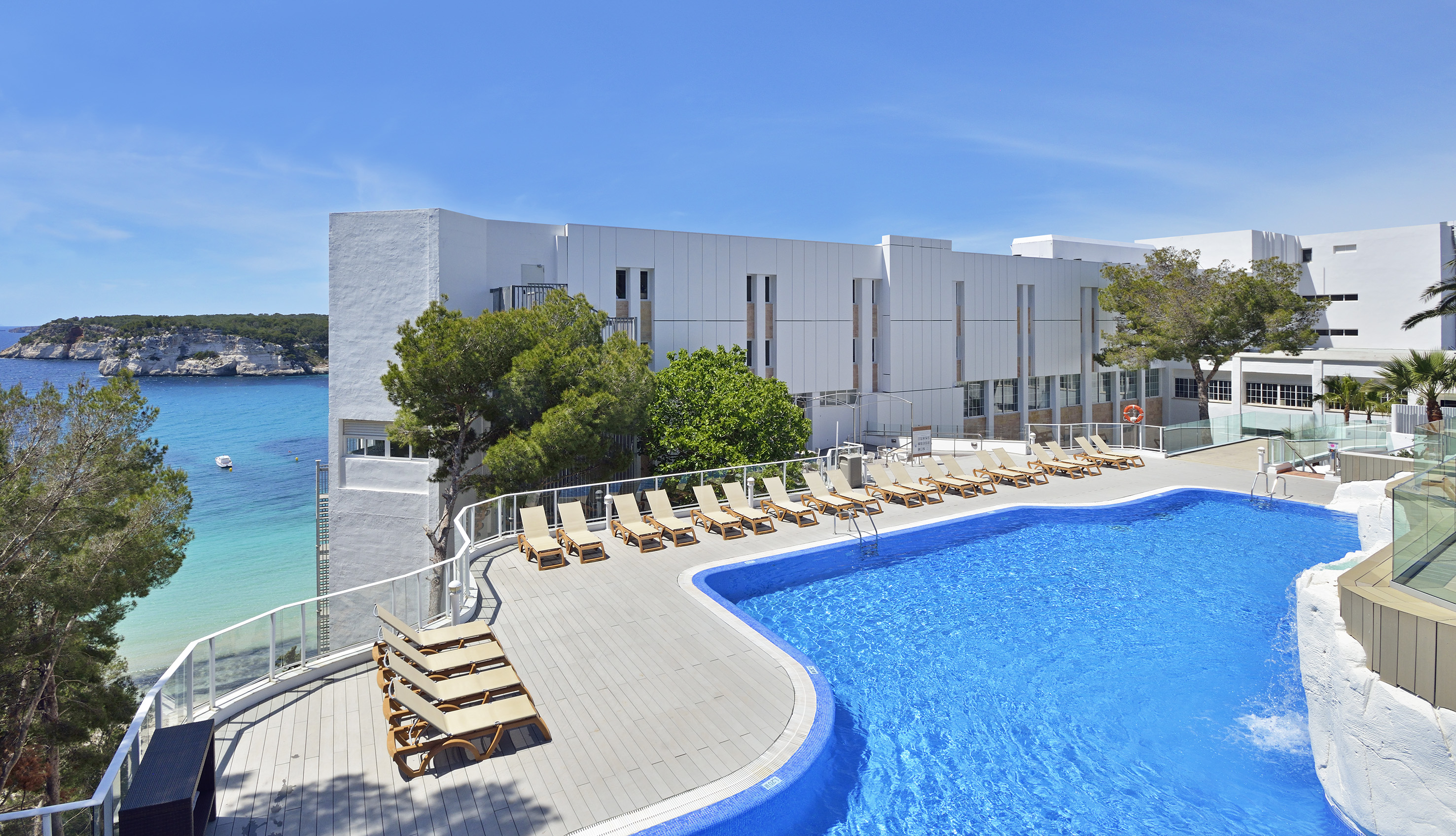 Hotel with swimming pool, sun loungers, overlooking turquoise sea and cliffs.