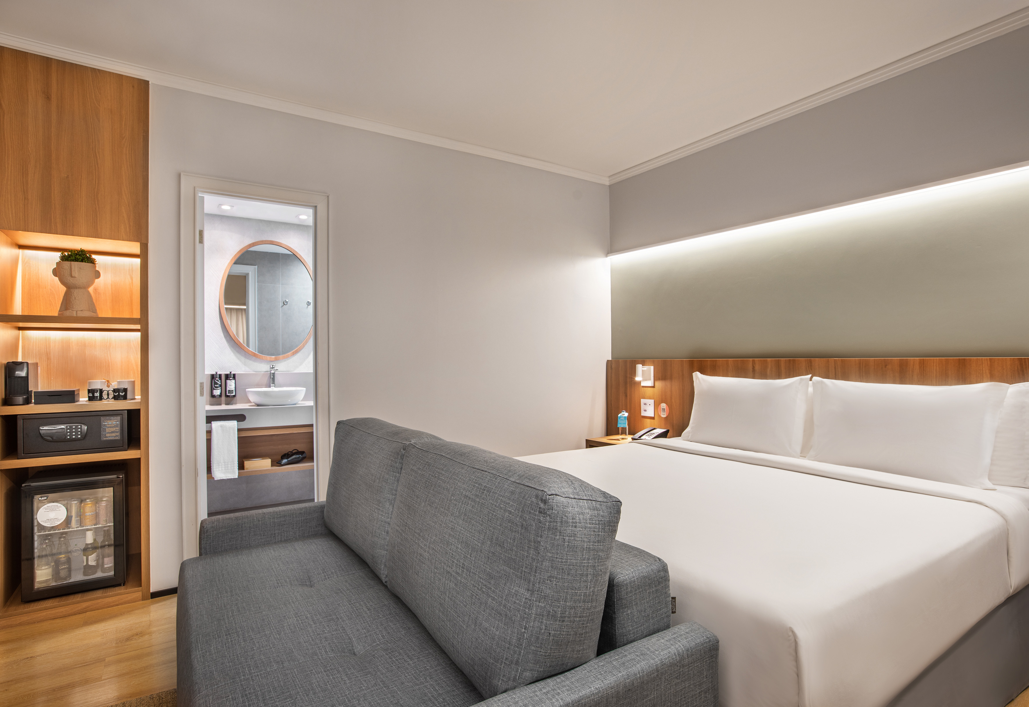 Modern hotel room with bed, gray sofa, mini-bar, and bathroom.