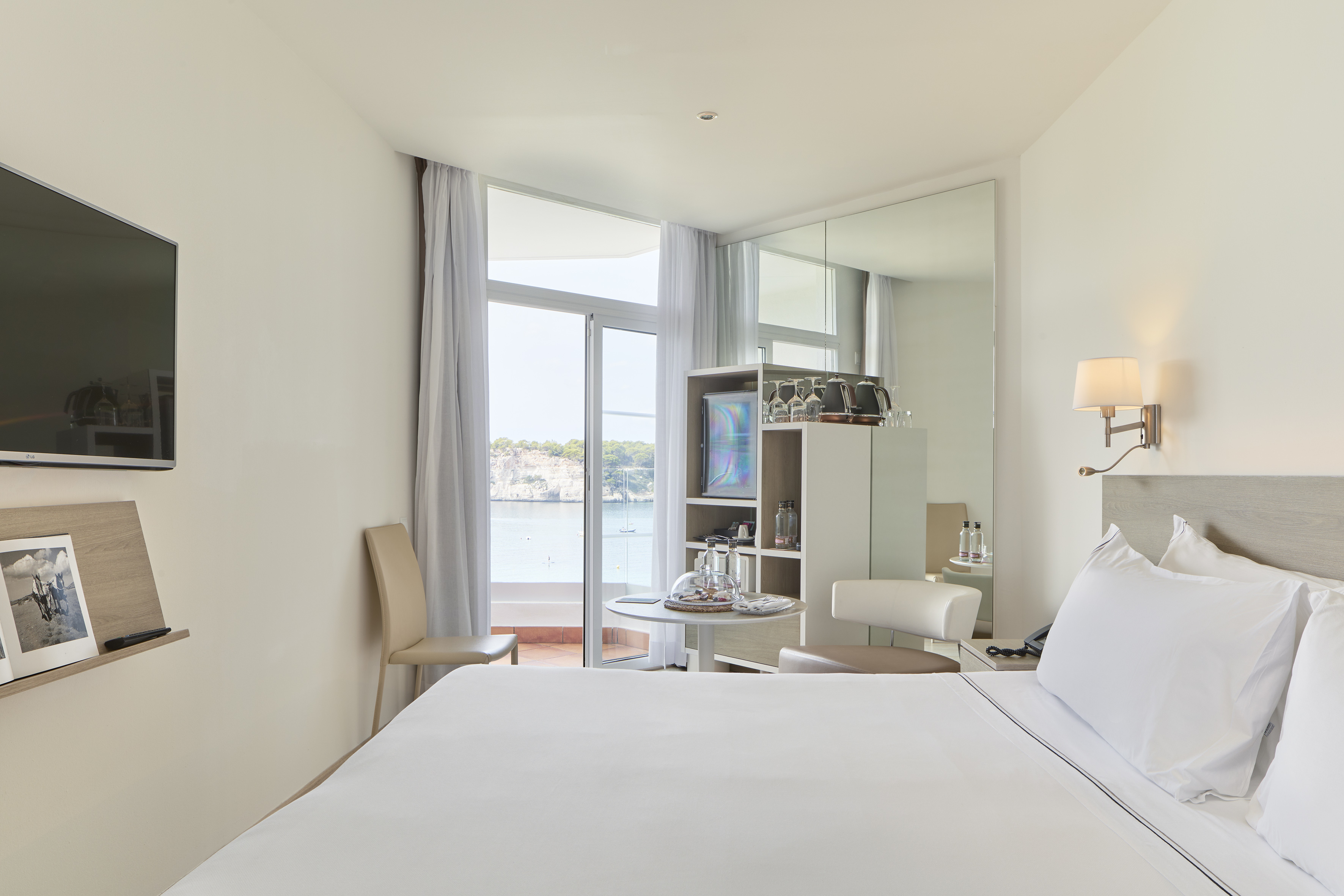 Bright hotel room with balcony, bay view, white bed, TV, and minibar.