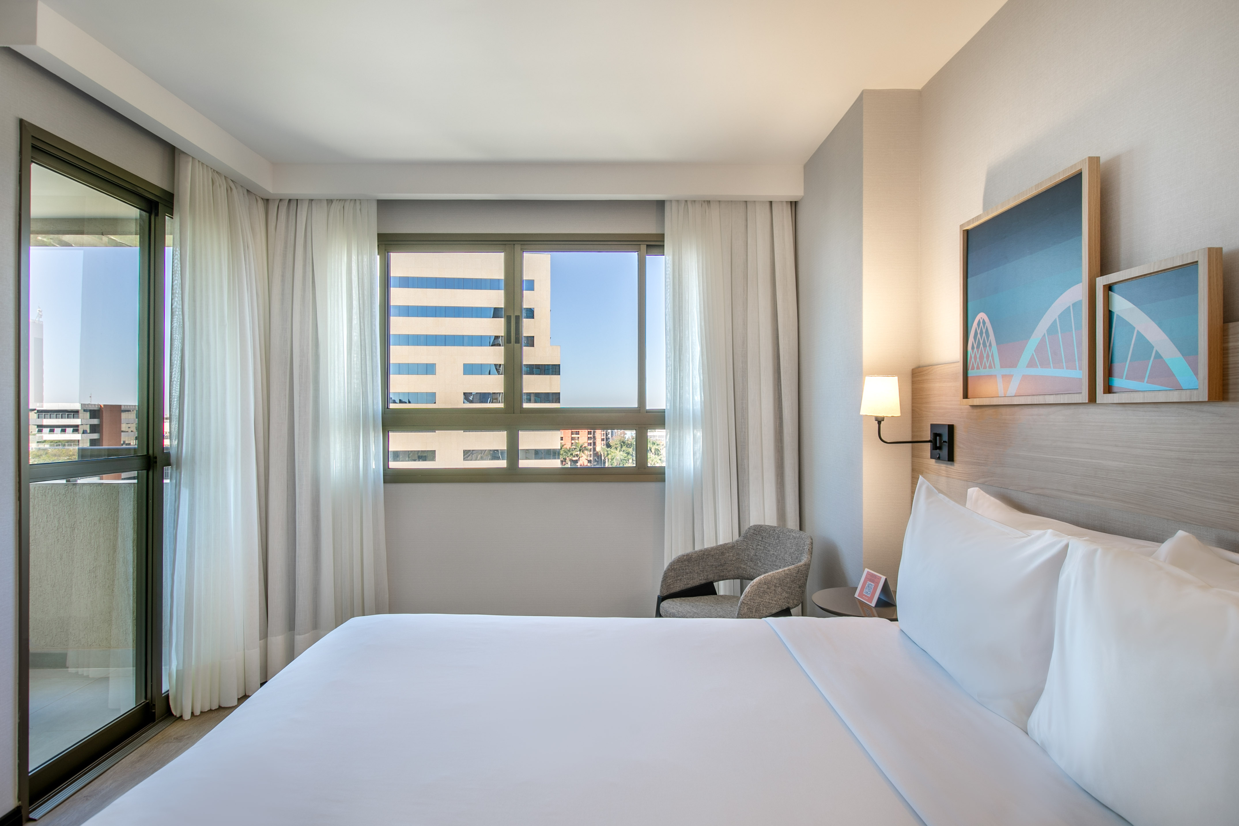 Modern hotel room with white bed, city view windows, sheer curtains, and abstract art.