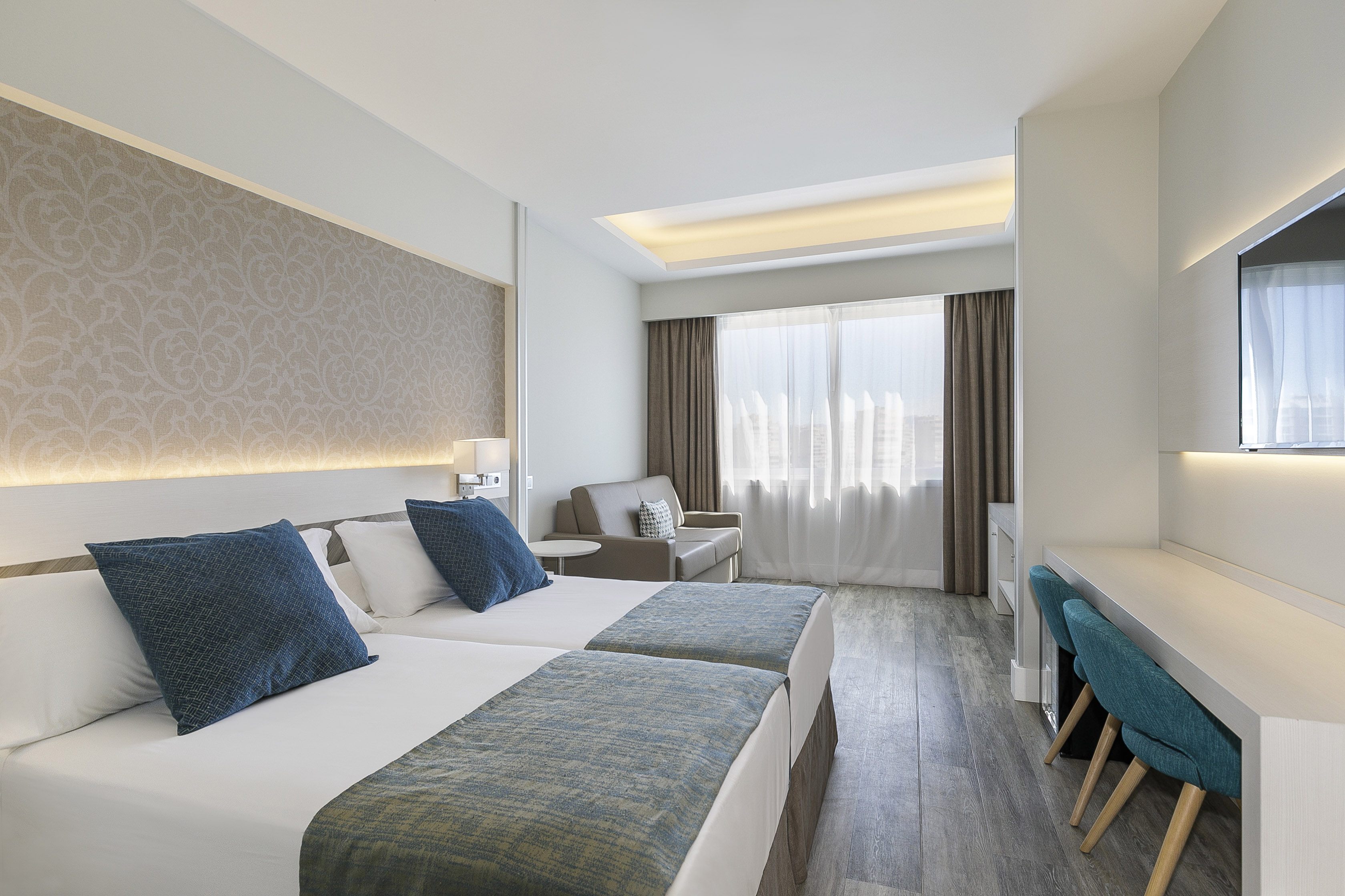 Modern hotel room with twin beds, sofa, desk, and bright window.