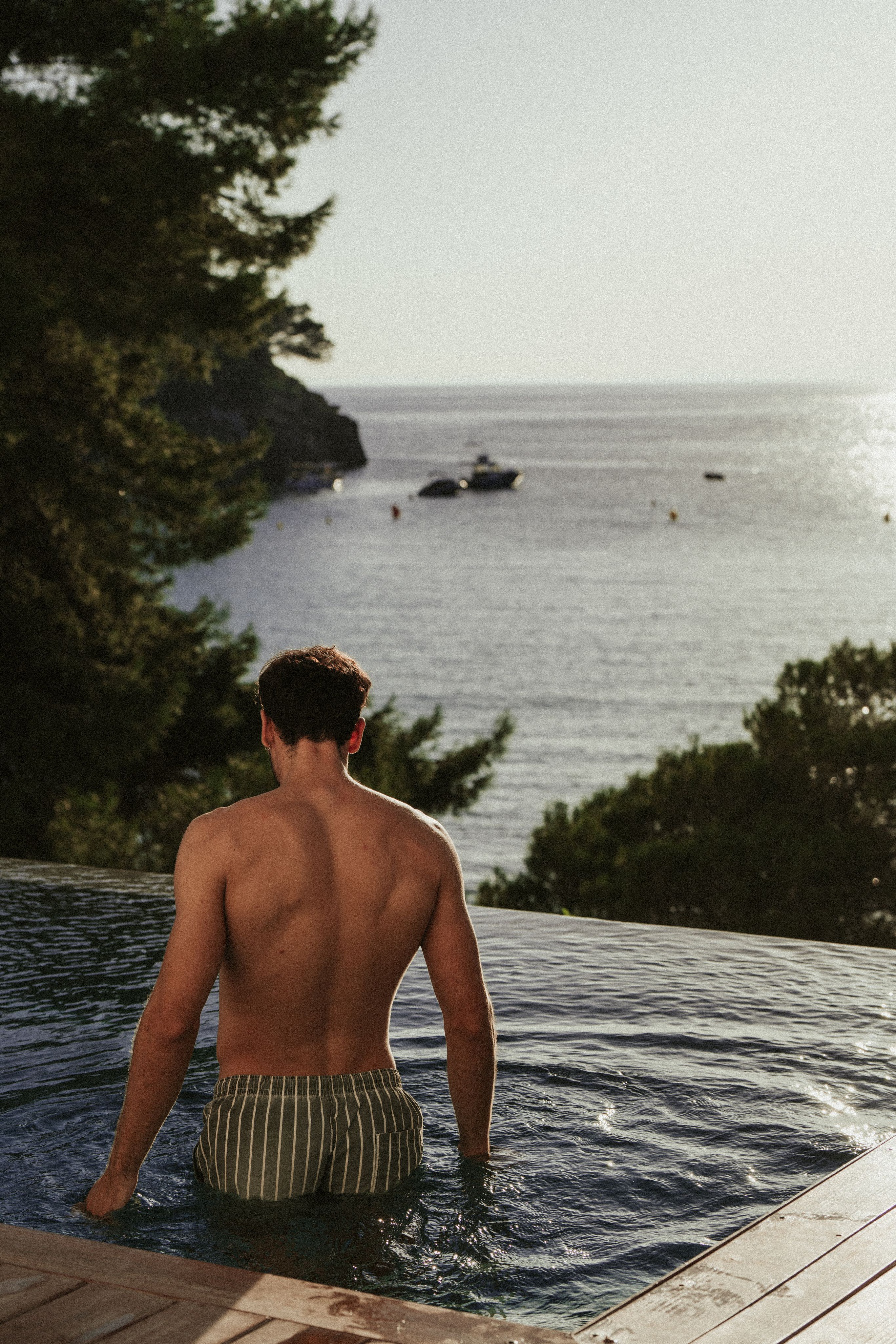 Man in striped trunks in infinity pool, looking at boats on ocean.