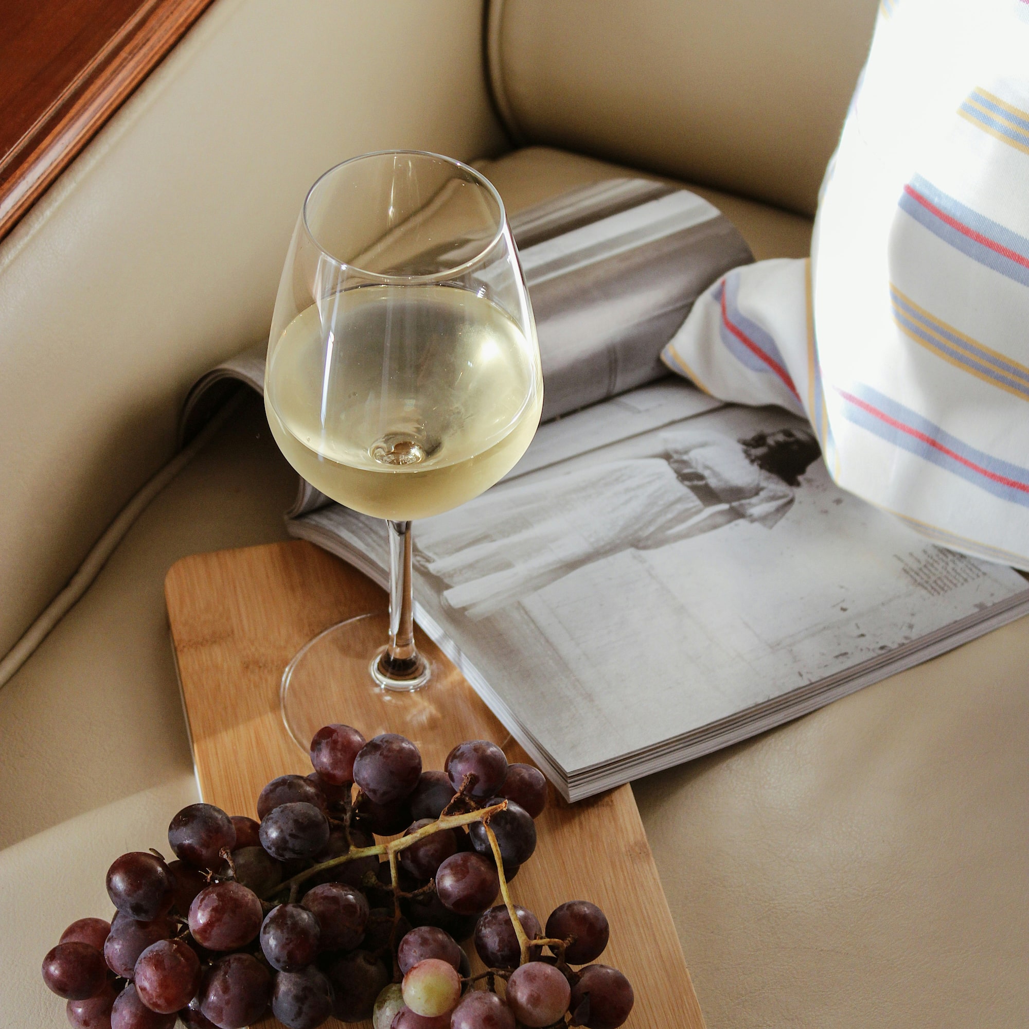 Glass of white wine, red grapes on wood board, open magazine, beige sofa.