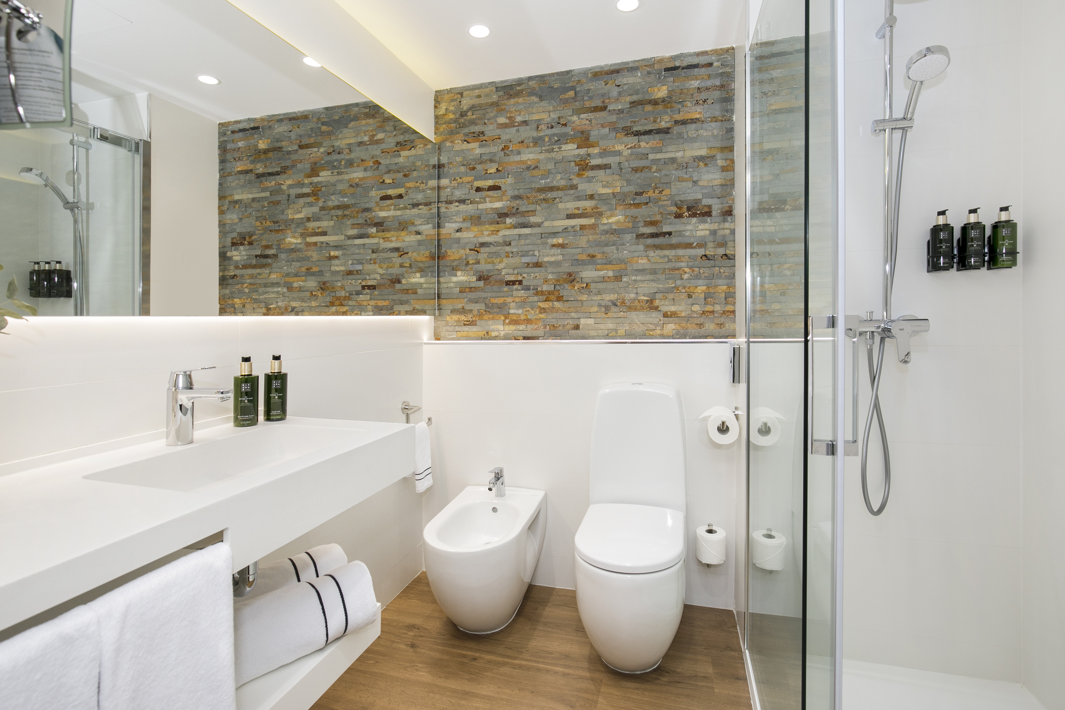 Modern bathroom: white vanity, toilet, bidet, glass shower, stone wall.