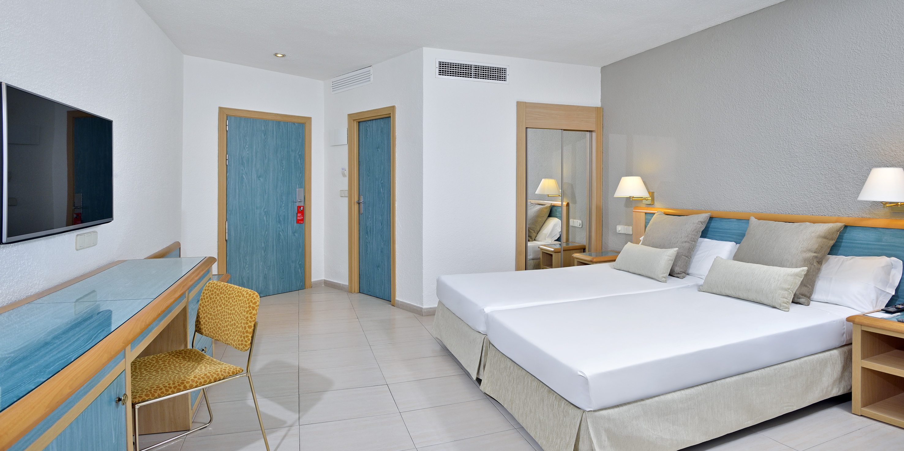 Bright modern hotel room with twin beds, blue doors, TV, and desk.