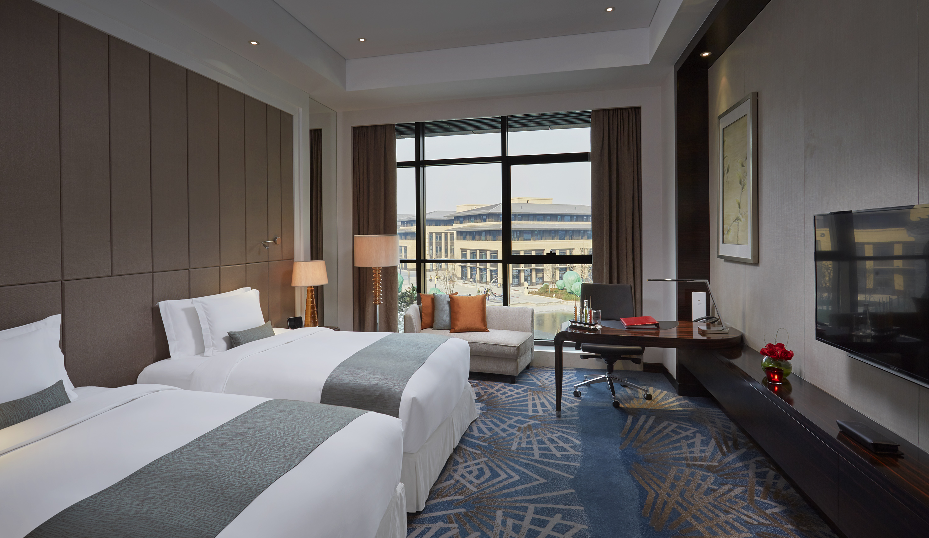Modern hotel room with two twin beds, desk, and city view from large window.