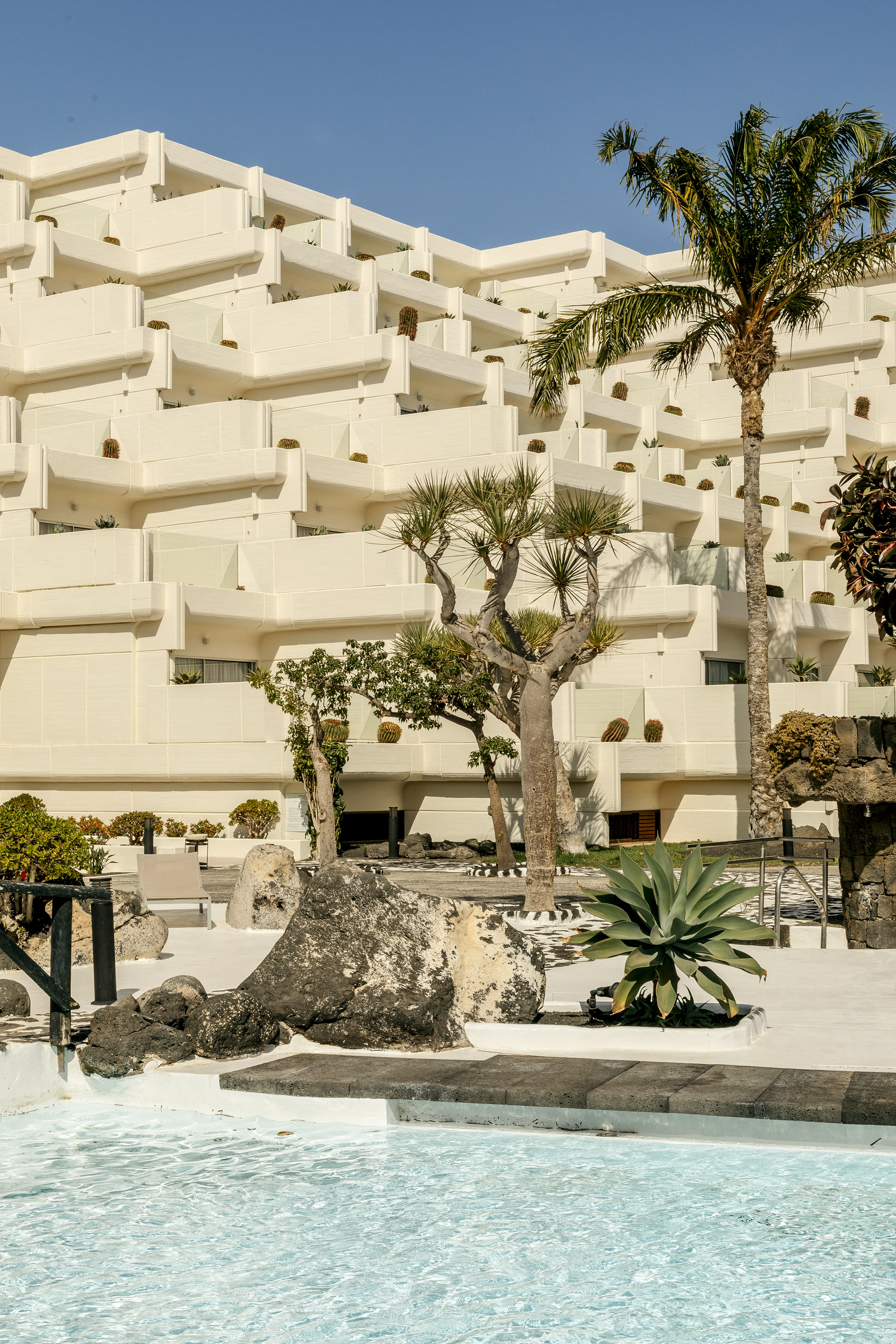 Sunny resort building with balconies, tropical trees, and a swimming pool.