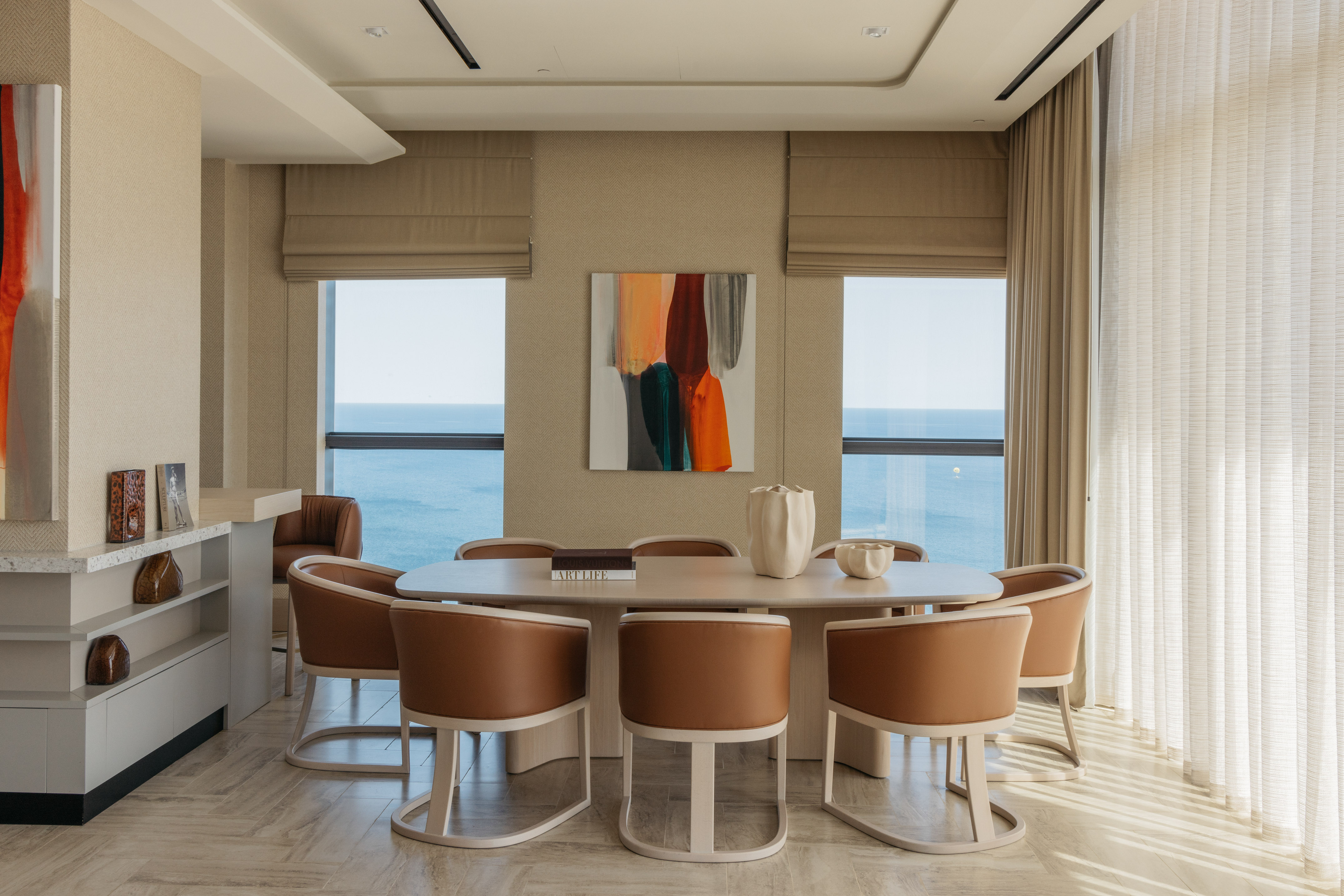 Modern dining room with ocean view, abstract art, and tan chairs.