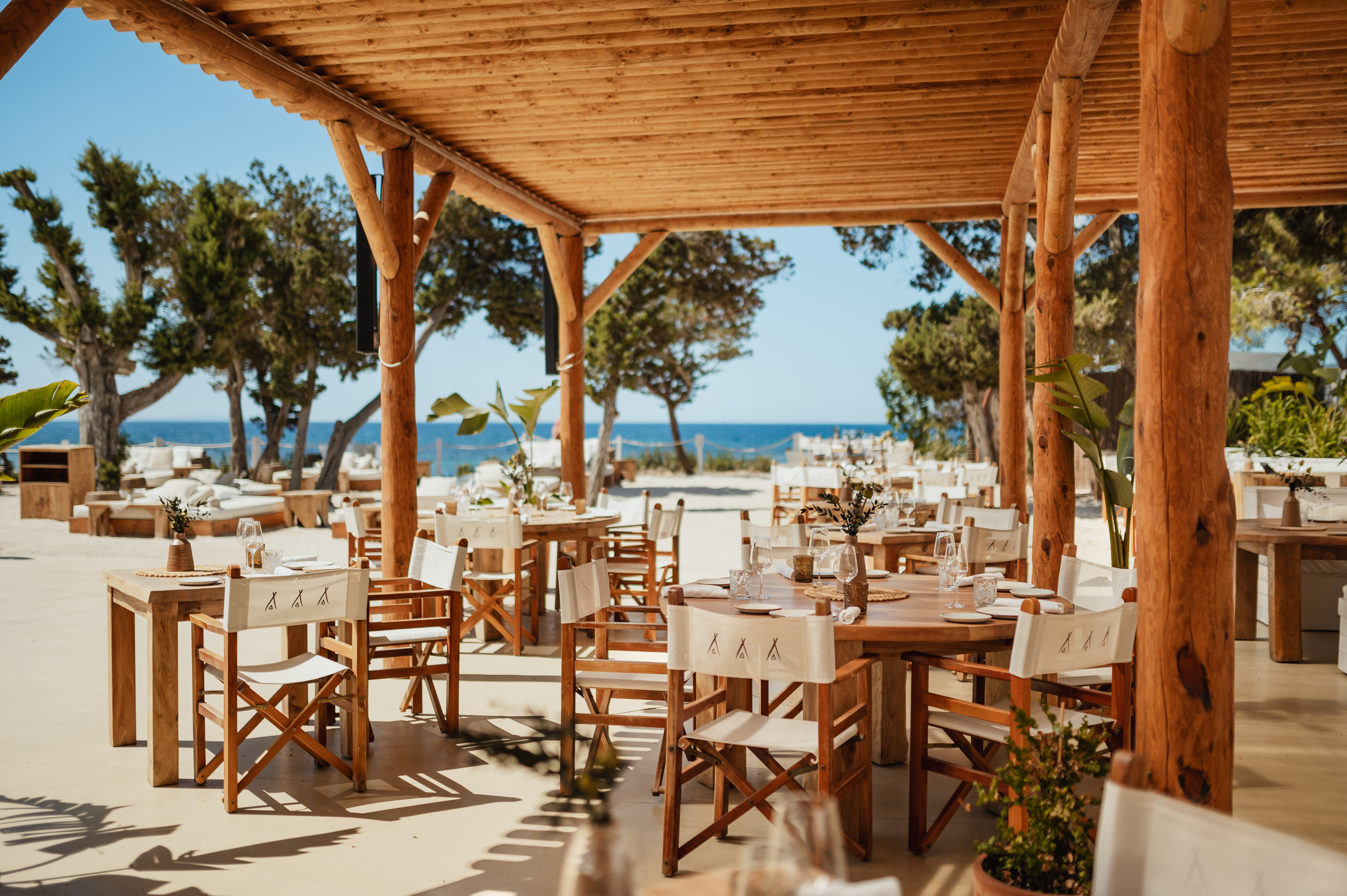 Beachfront restaurant under wooden pergola, outdoor dining tables, and blue sea.
