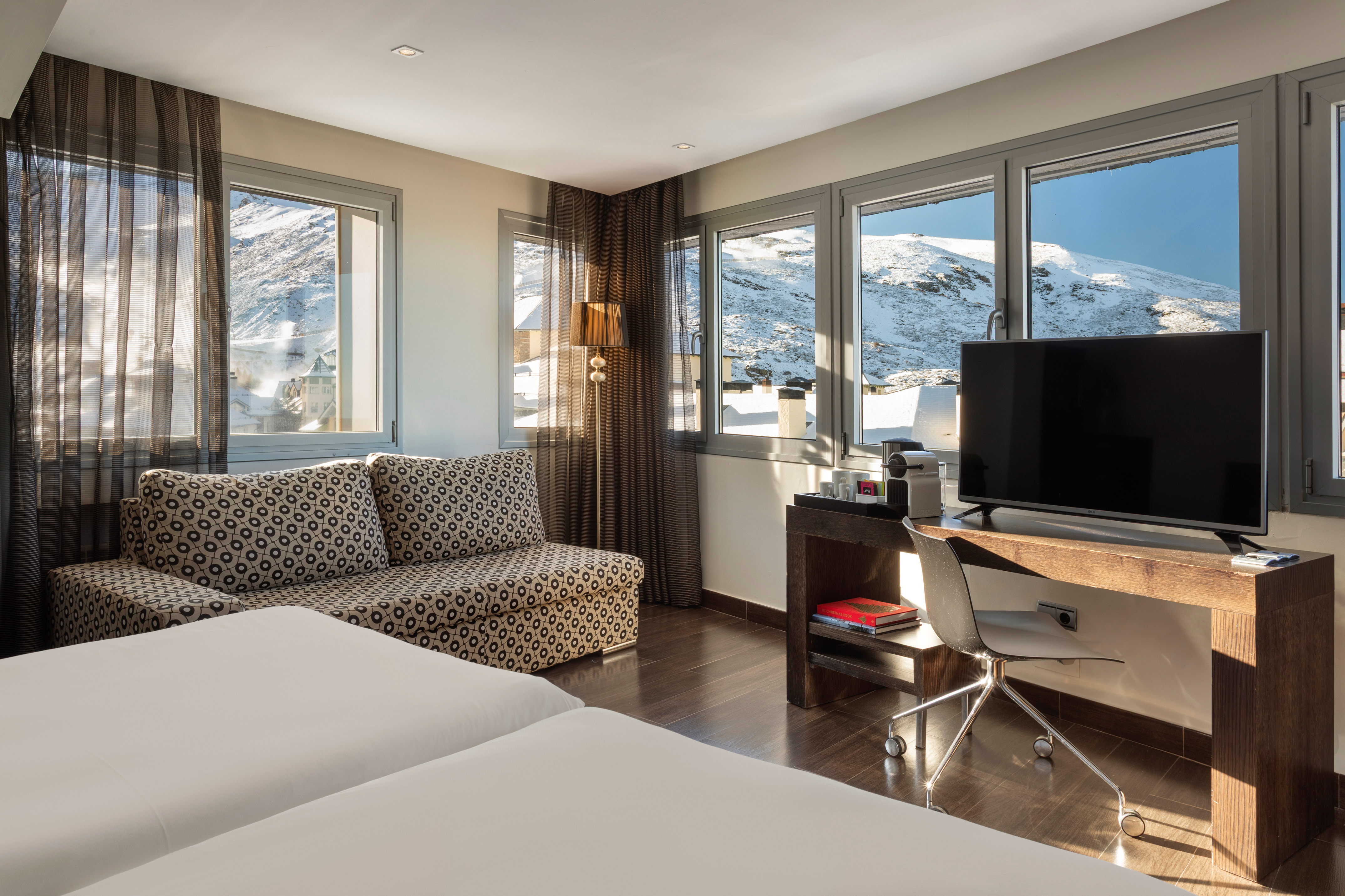 Hotel room with snowy mountain view, patterned sofa, desk, and TV.