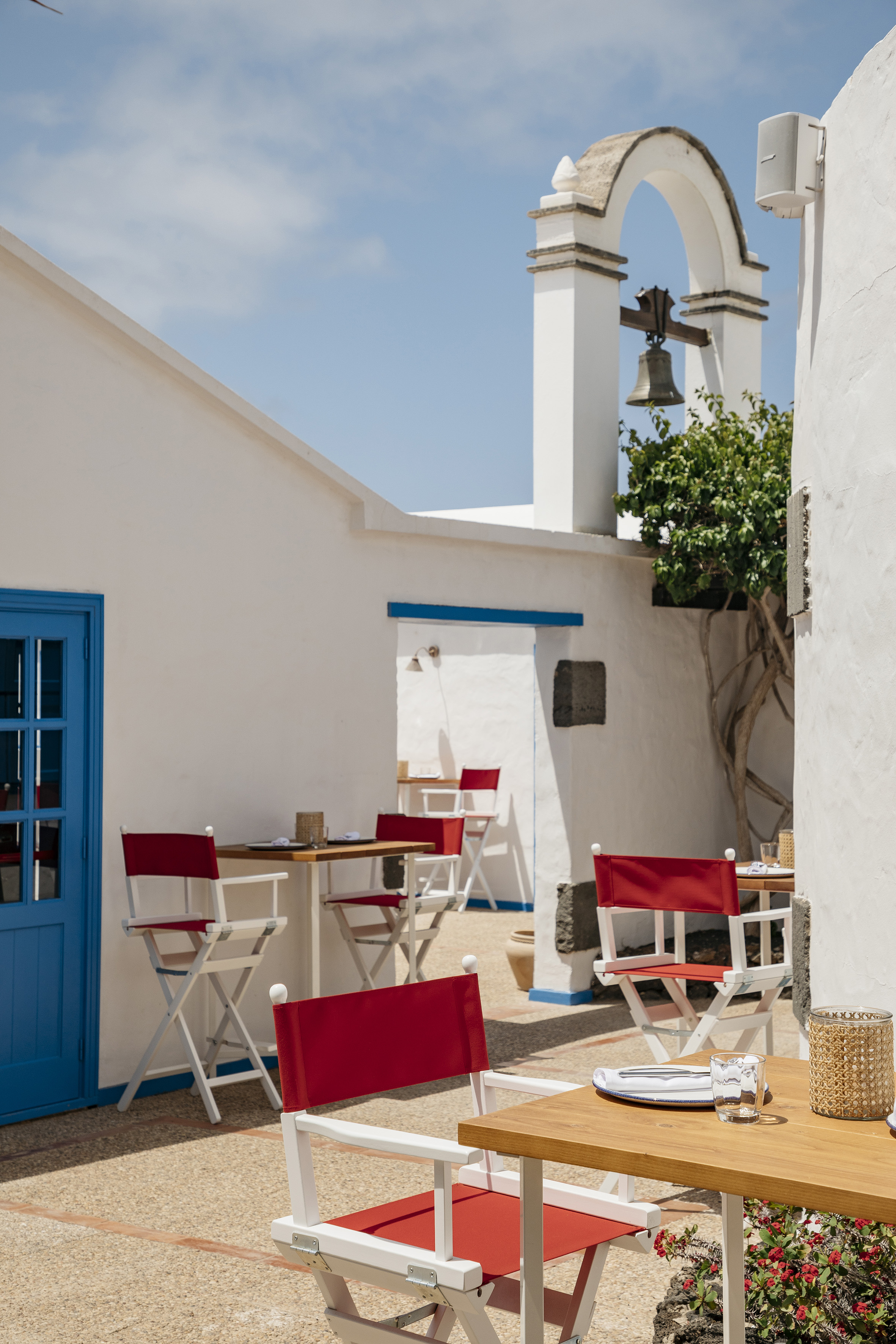 White Mediterranean restaurant patio, blue door, red chairs, bell tower.