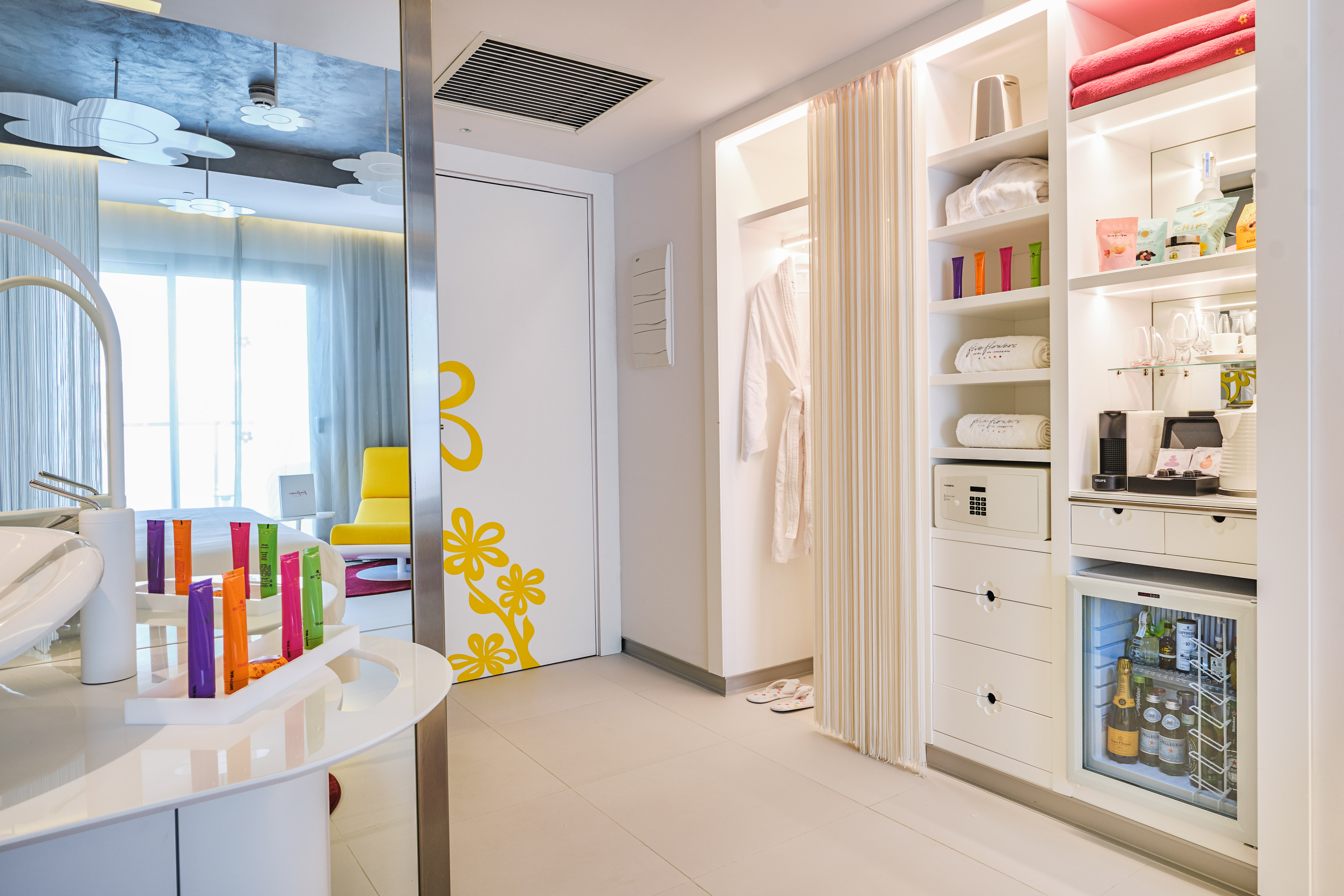 Bright hotel room entrance with white wardrobe, stocked minibar, and safe.