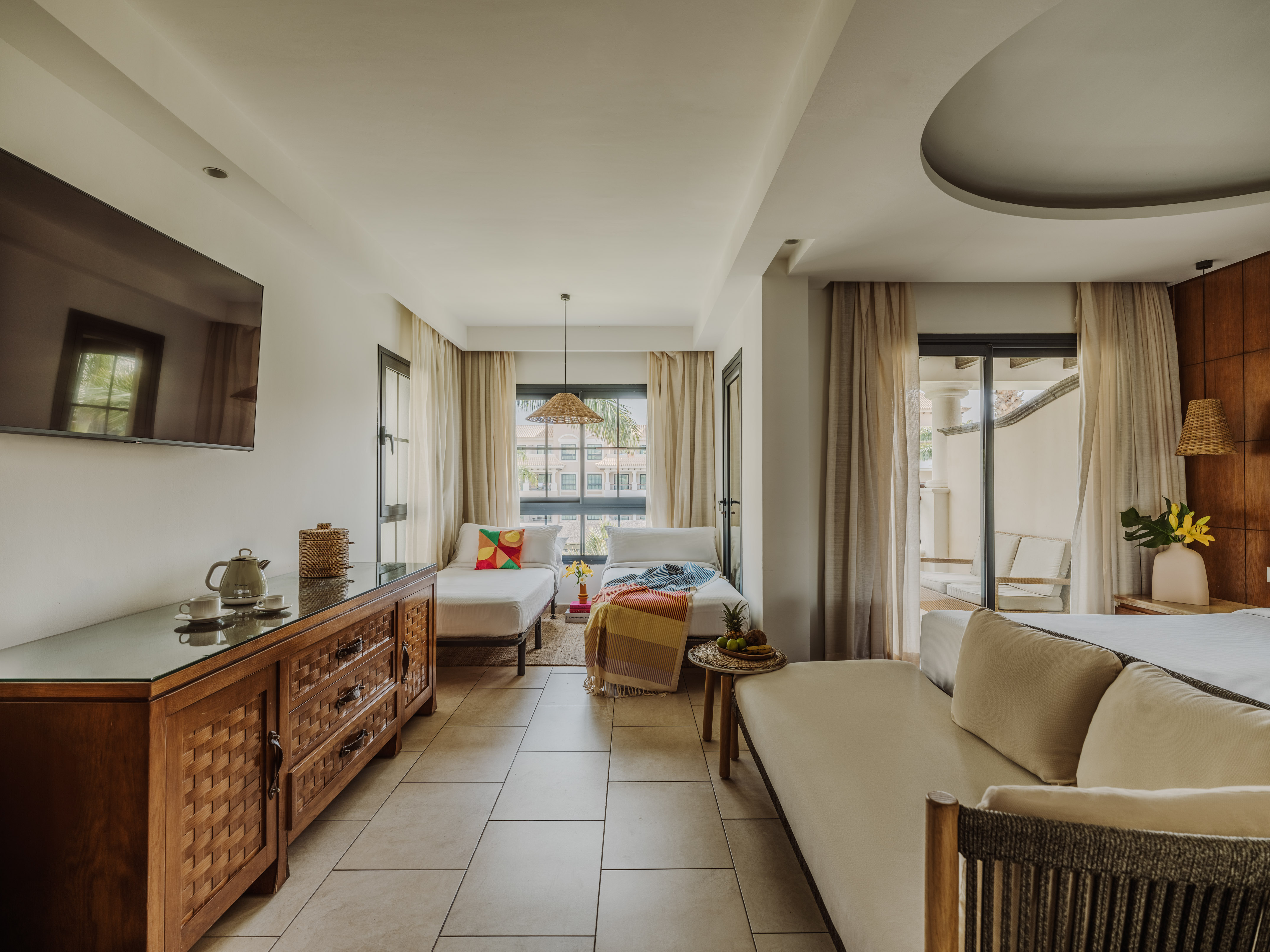 Stylish hotel suite with daybeds, large TV, wooden dresser, and balcony view.
