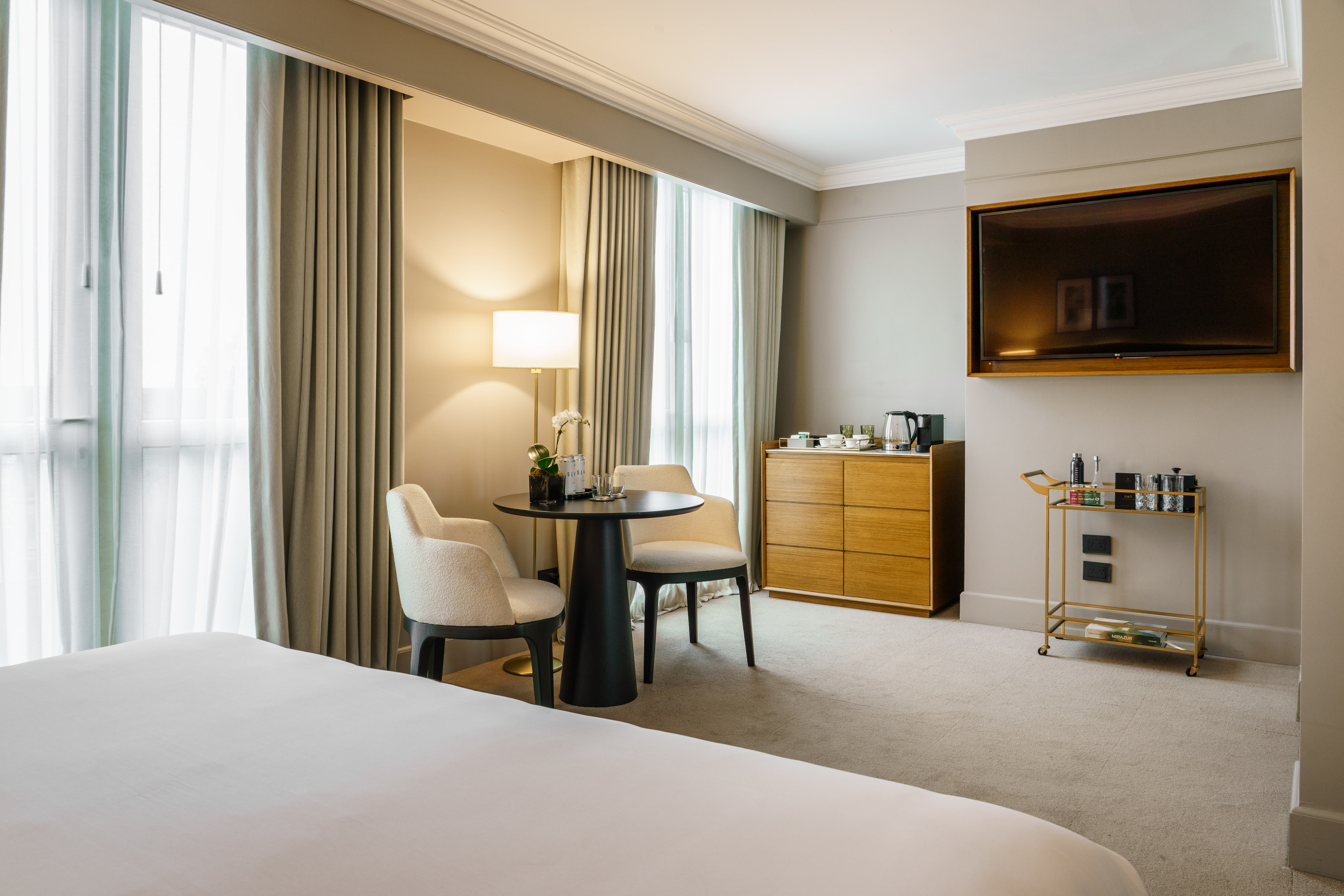 Modern hotel room with bed, stylish sitting area, wall TV, and bar cart.